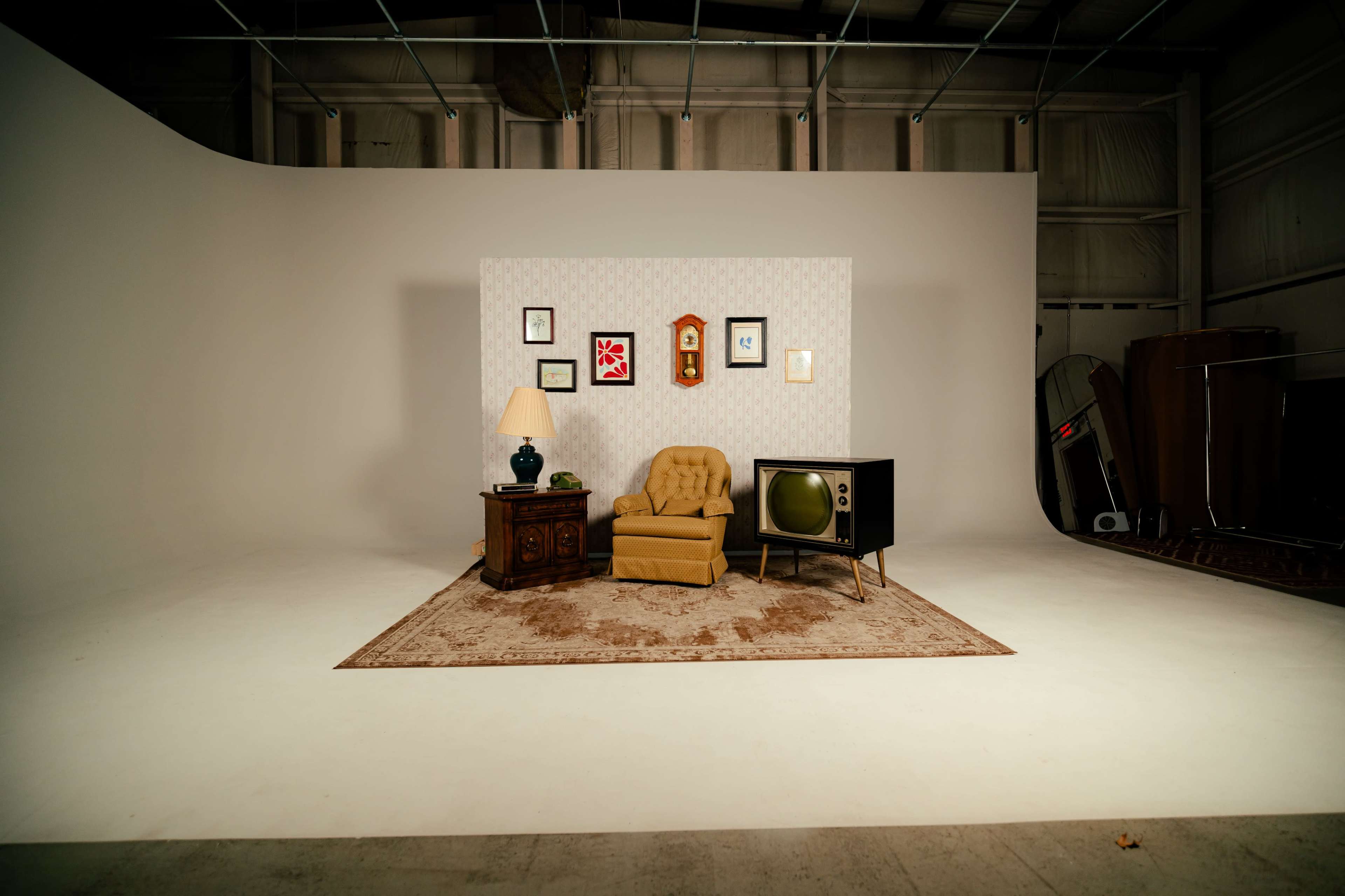 The image shows a vintage-inspired living room setup featuring a yellow armchair, a side table with a lamp, a retro television, and framed pictures on a patterned wall, all placed on a large area rug in a spacious studio.