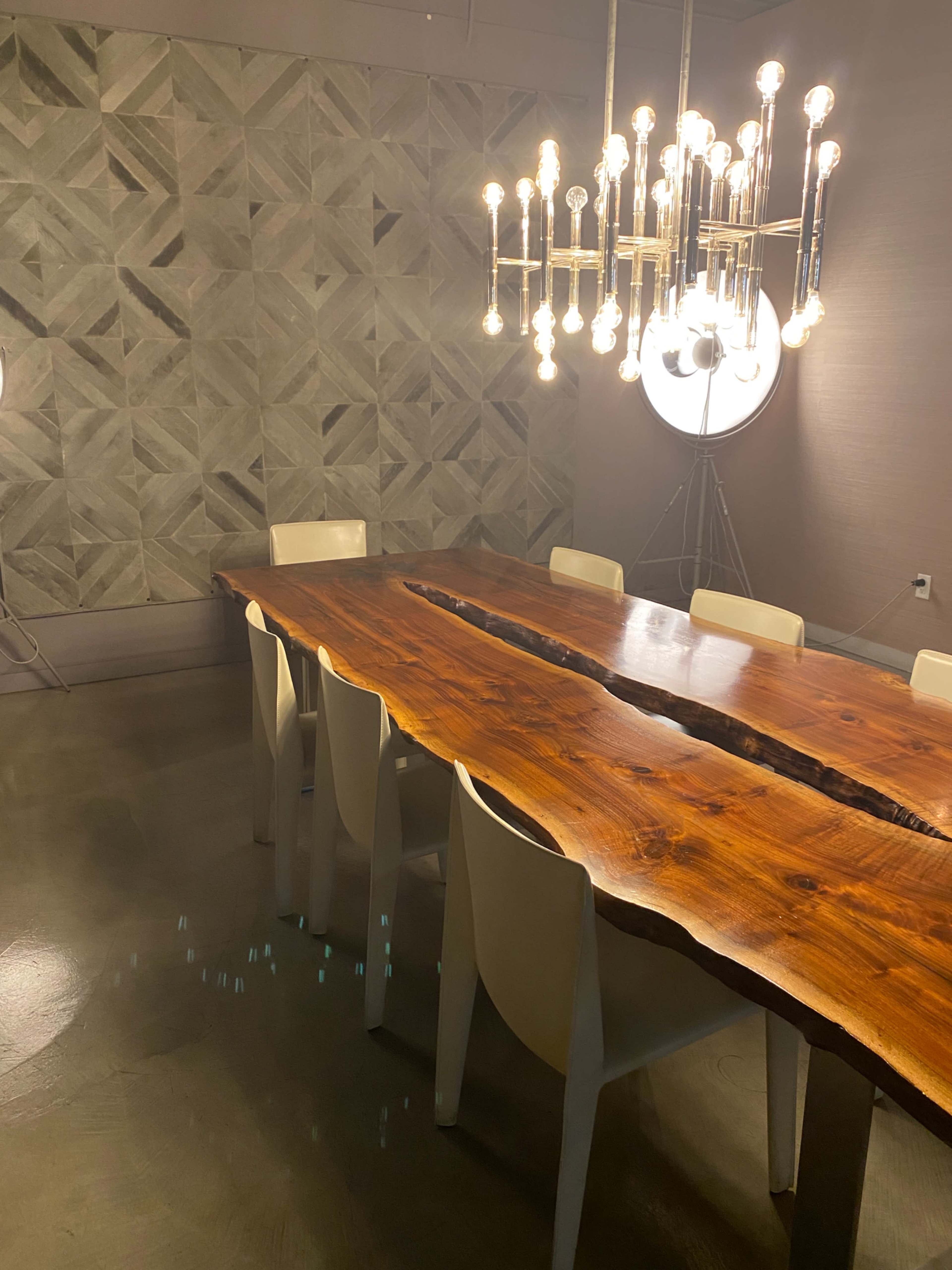 A long wooden table with a natural edge is situated in a modern meeting room, surrounded by white chairs, with a geometric-patterned wall and a distinctive chandelier overhead.