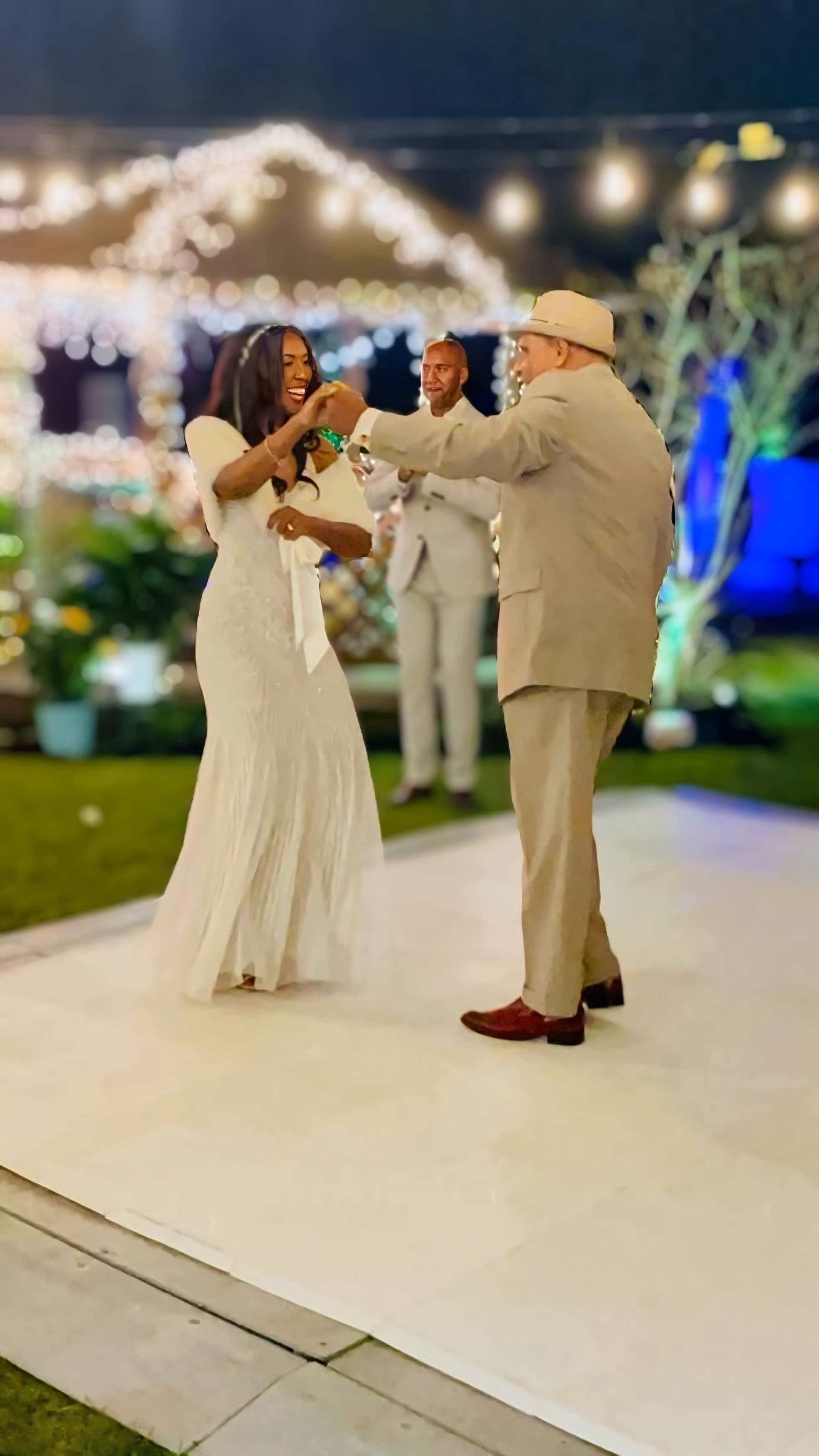 A bride in a white dress dances joyfully while holding hands with a man in a suit, with a second man looking on at a festive outdoor setting.