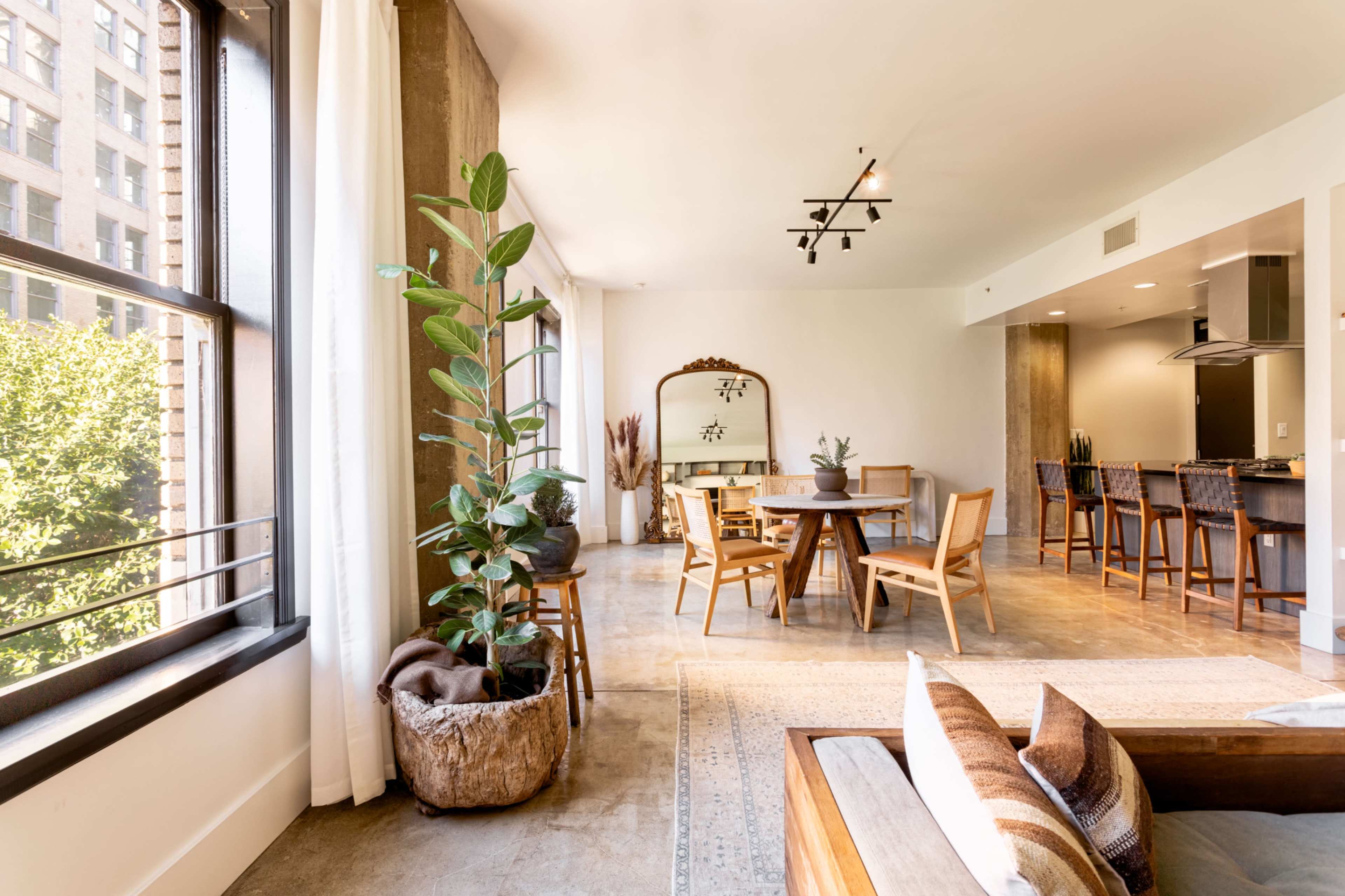 A spacious living area features a dining table, wooden chairs, large windows, and a kitchen bar with high stools.