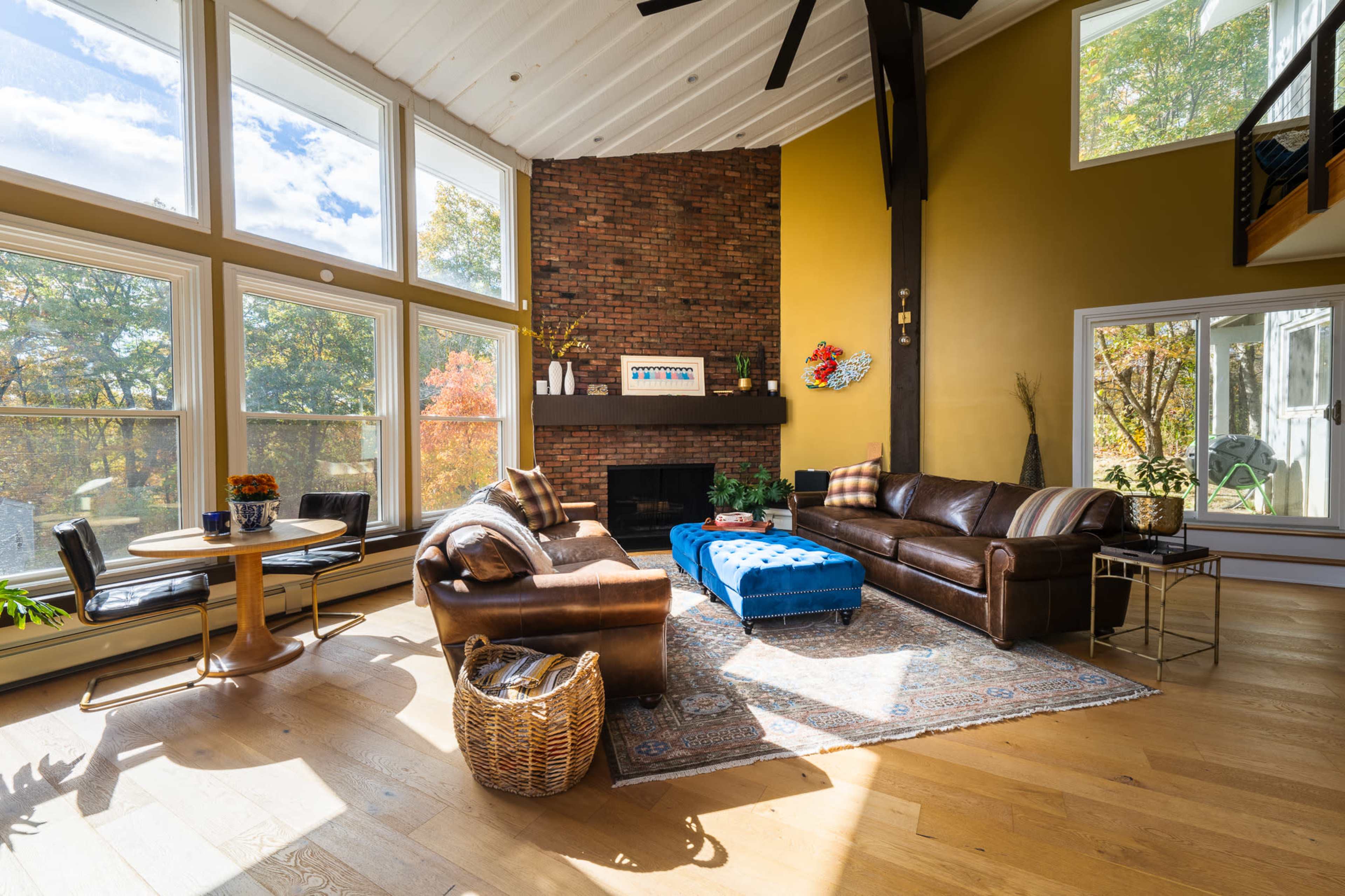 The image shows a spacious living room with large windows, a brown leather sofa, a blue ottoman, and a dining table set near a fireplace surrounded by autumn foliage outside.