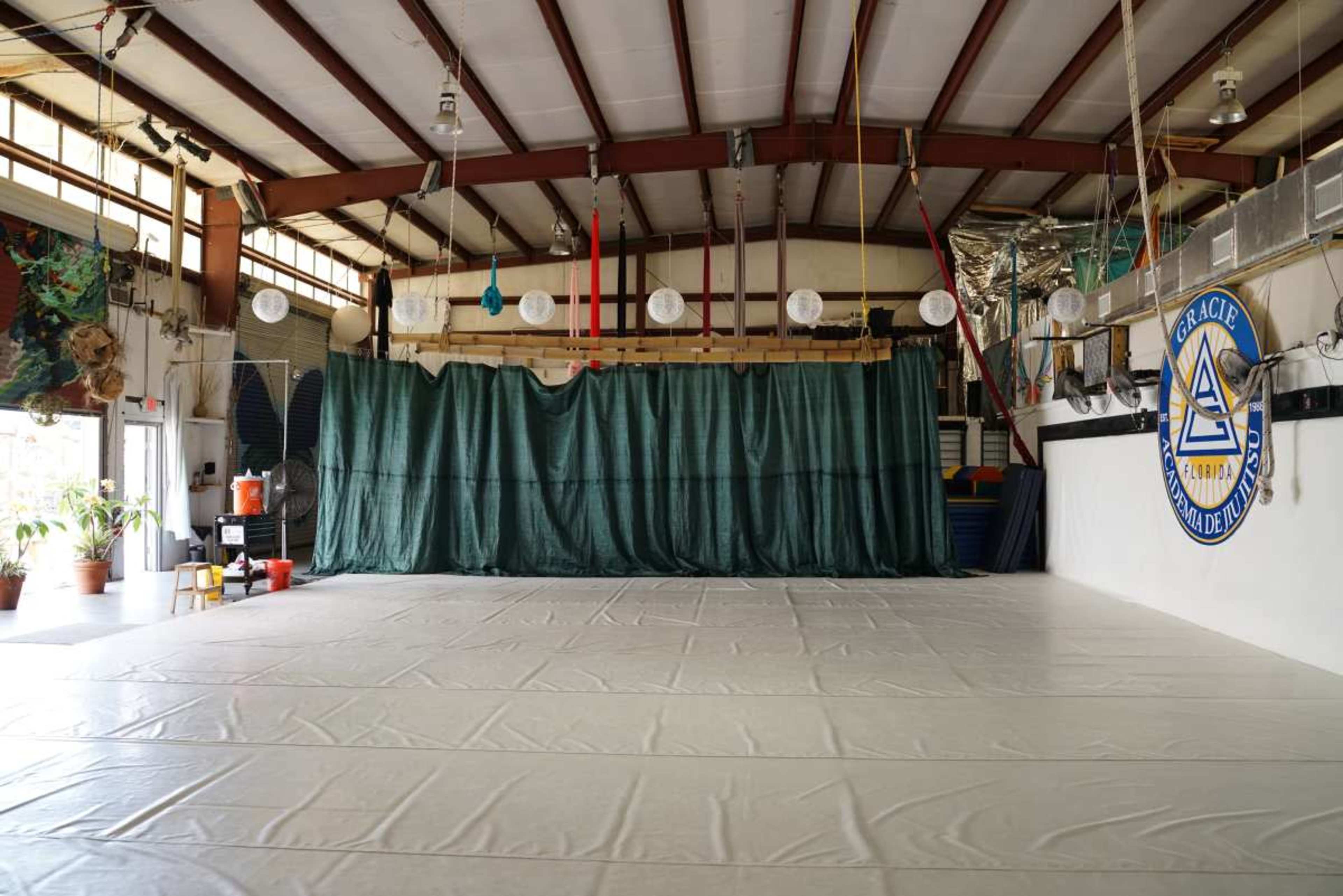 The image shows a spacious indoor gymnasium with a large, empty mat area and green curtains hanging at the back.