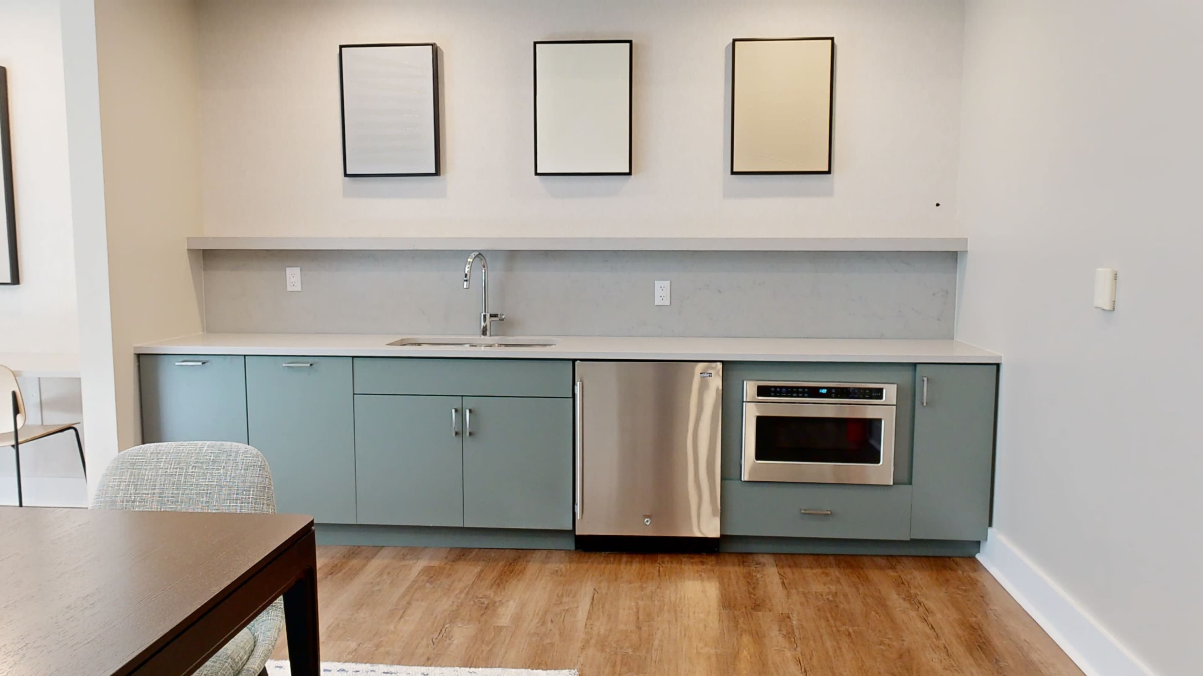A modern kitchen with light green cabinets, a stainless steel dishwasher and microwave, and three empty framed pictures on the wall above the countertop.