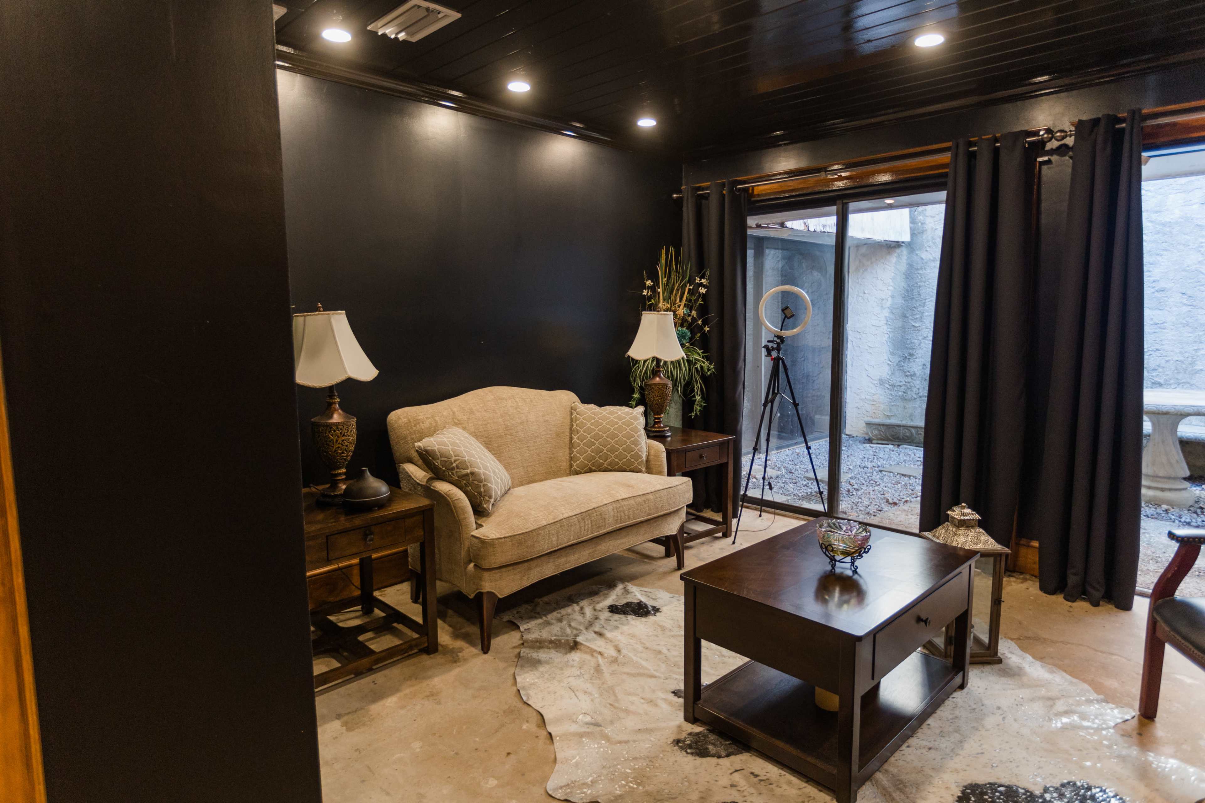 A cozy room with dark walls, a beige sofa, two lamps on side tables, and a coffee table on a stained floor, leading to a window with gray curtains.