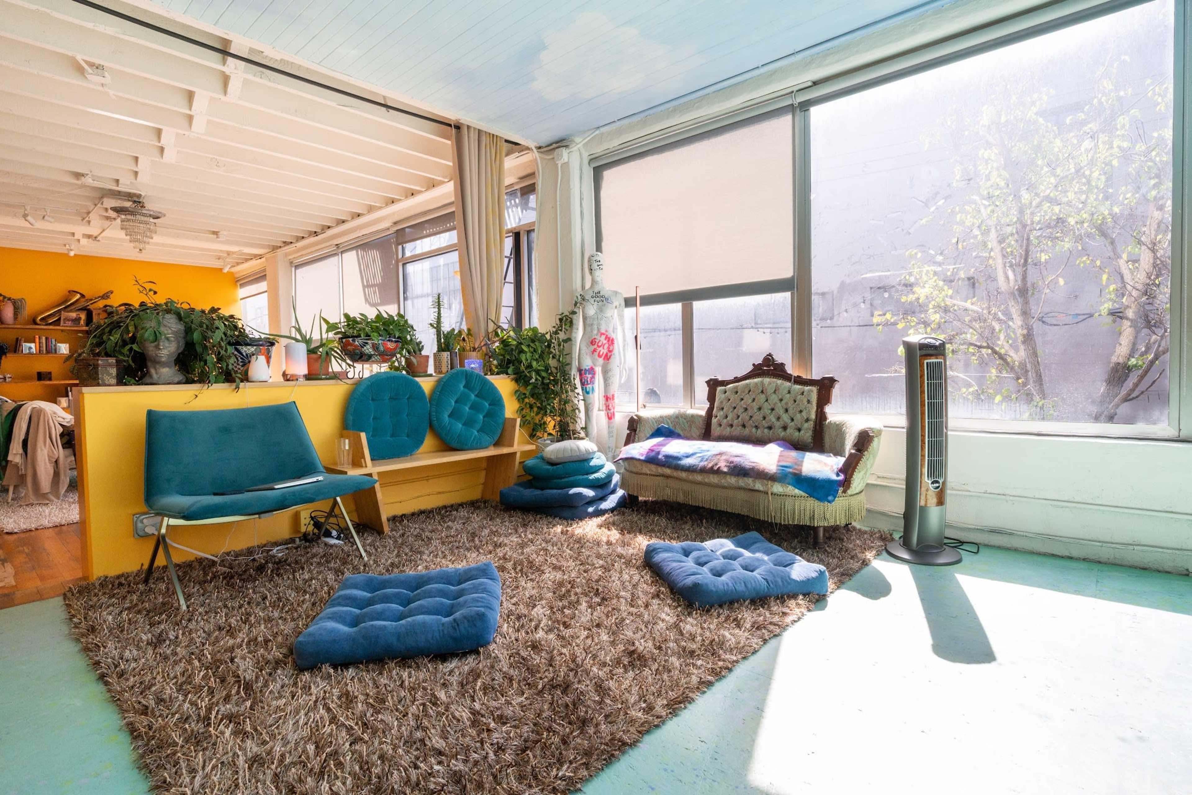 The image shows a bright, open space featuring a comfortable seating area with blue cushions, a vintage sofa, and large windows allowing natural light to fill the room.