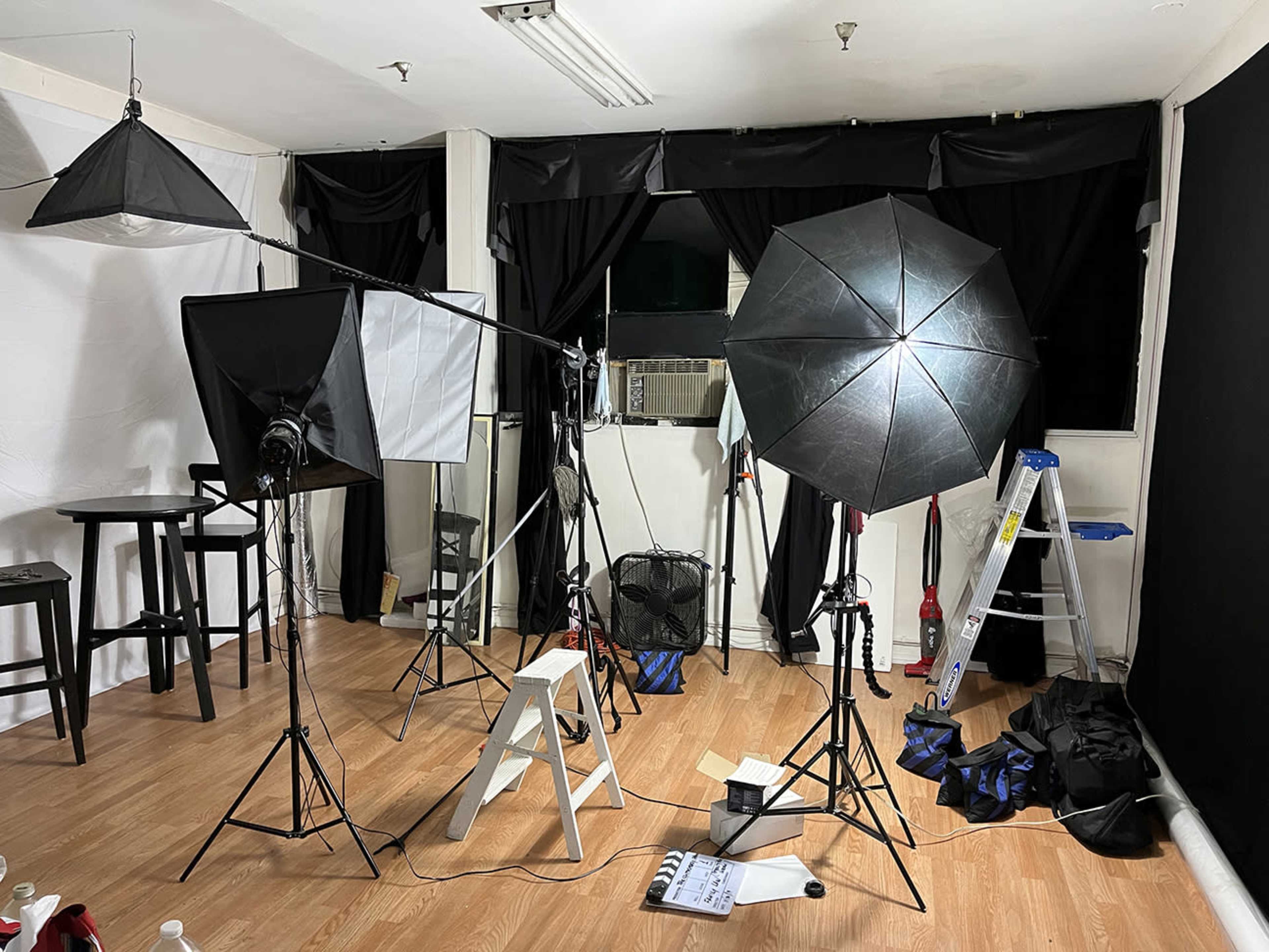 A photography studio setup with multiple light stands, softboxes, and a backdrop, featuring wooden flooring and various equipment arranged throughout the space.