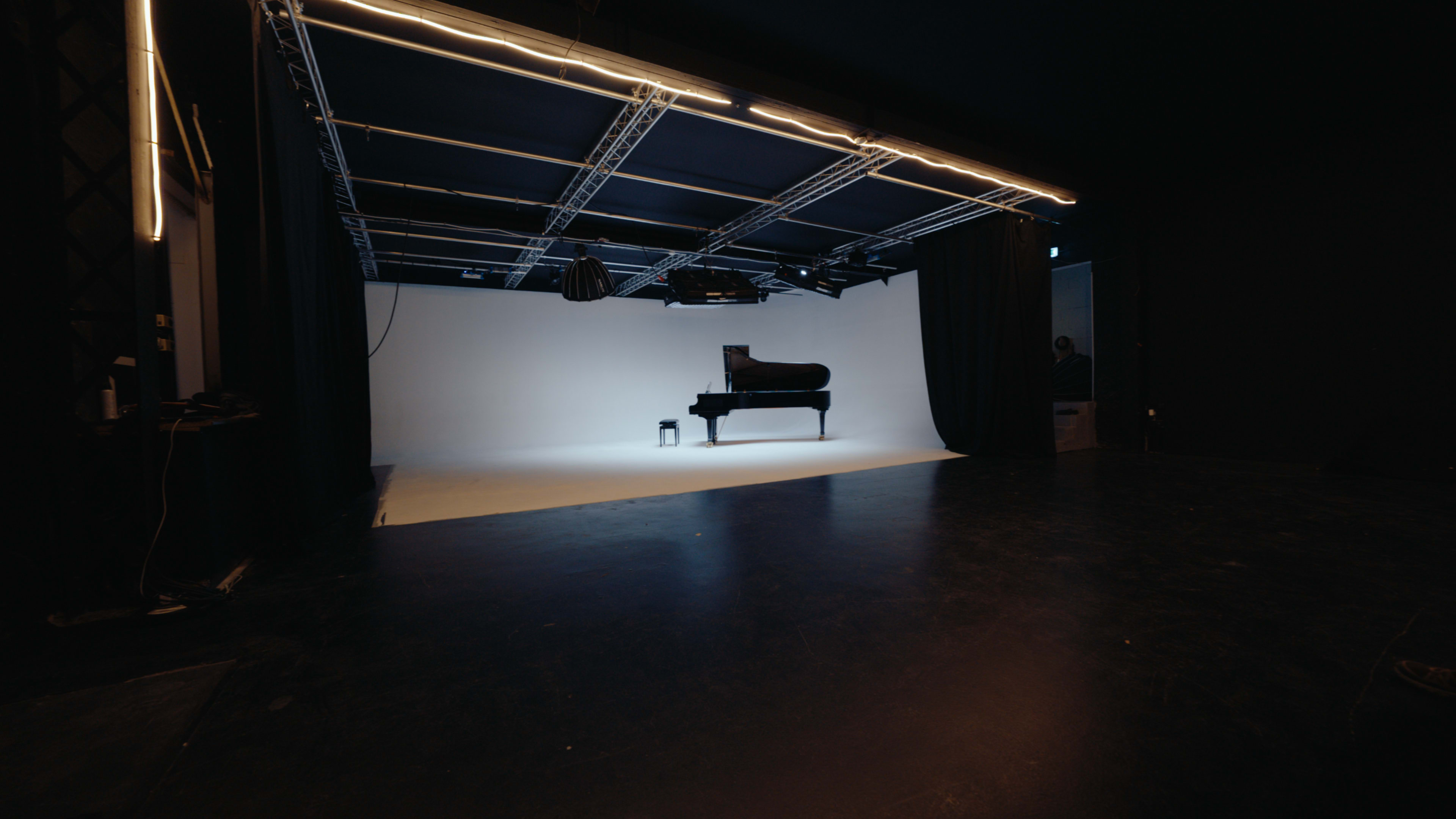 The image shows a dimly lit studio space with a grand piano sitting against a white backdrop, illuminated by overhead lights.