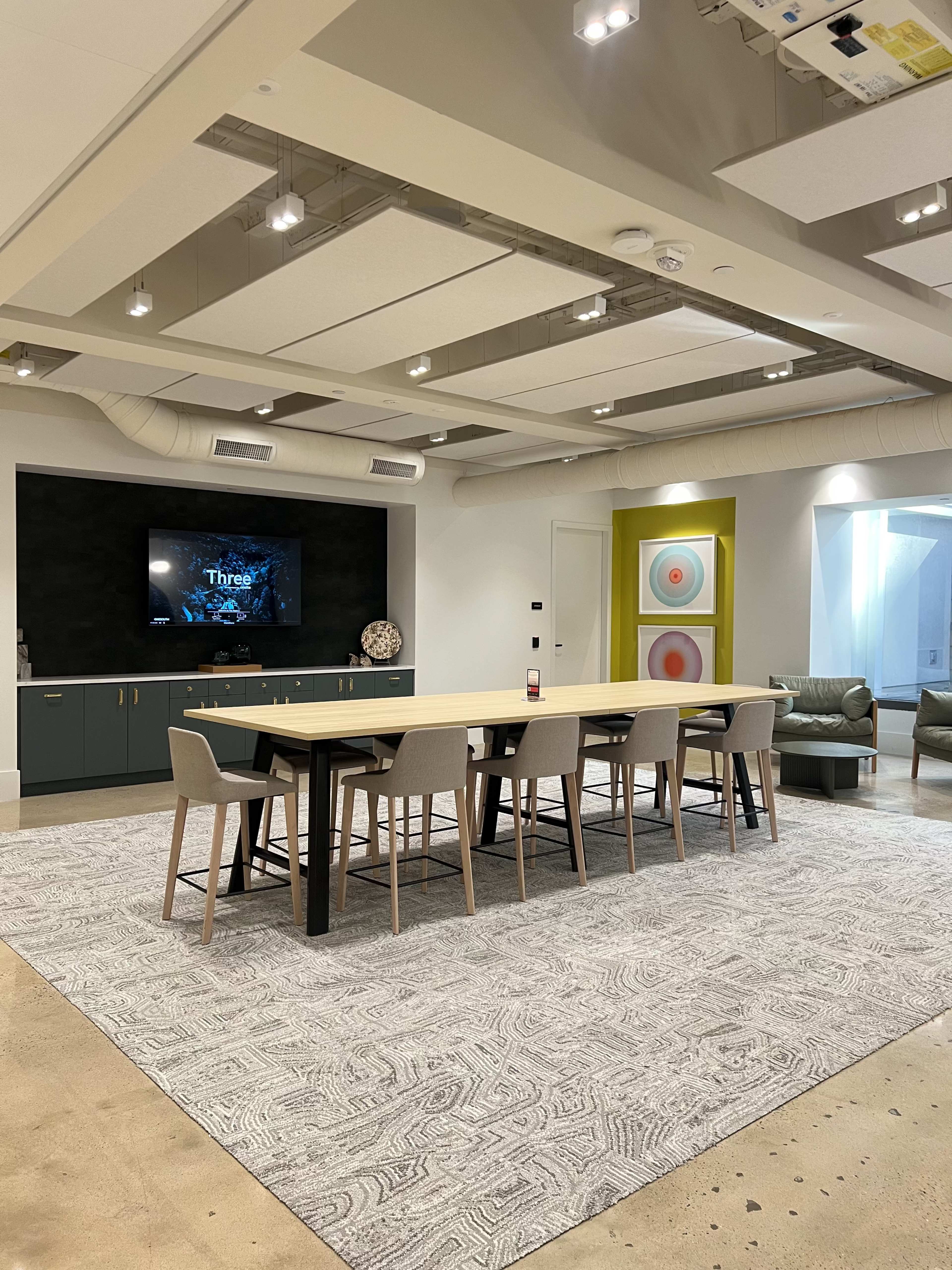 A modern conference room features a large table surrounded by chairs, a wall-mounted screen, and a patterned rug on the floor.