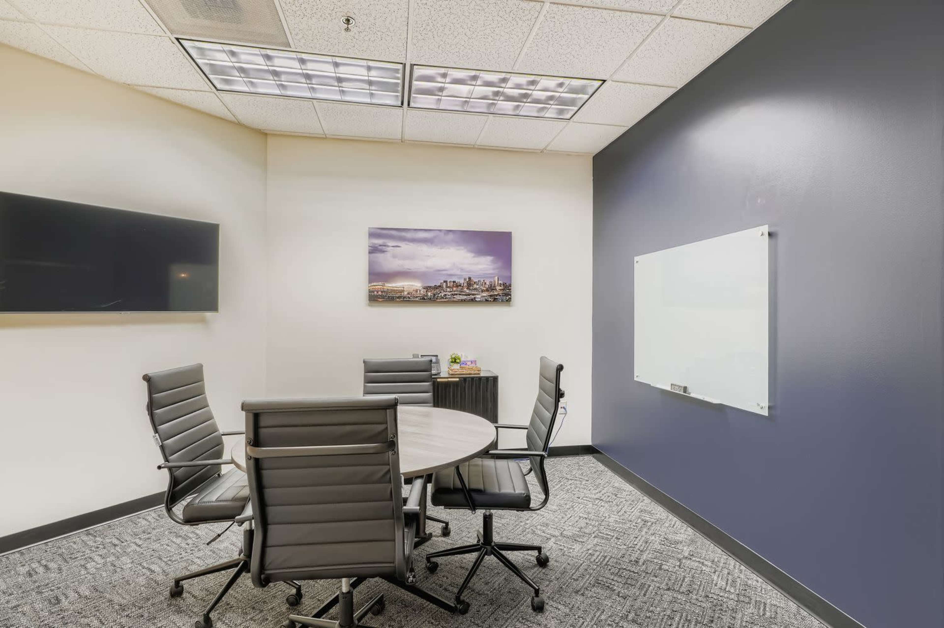 The image shows a small conference room with a round table and four black chairs, a wall-mounted television, a whiteboard, and a cityscape picture on the wall.