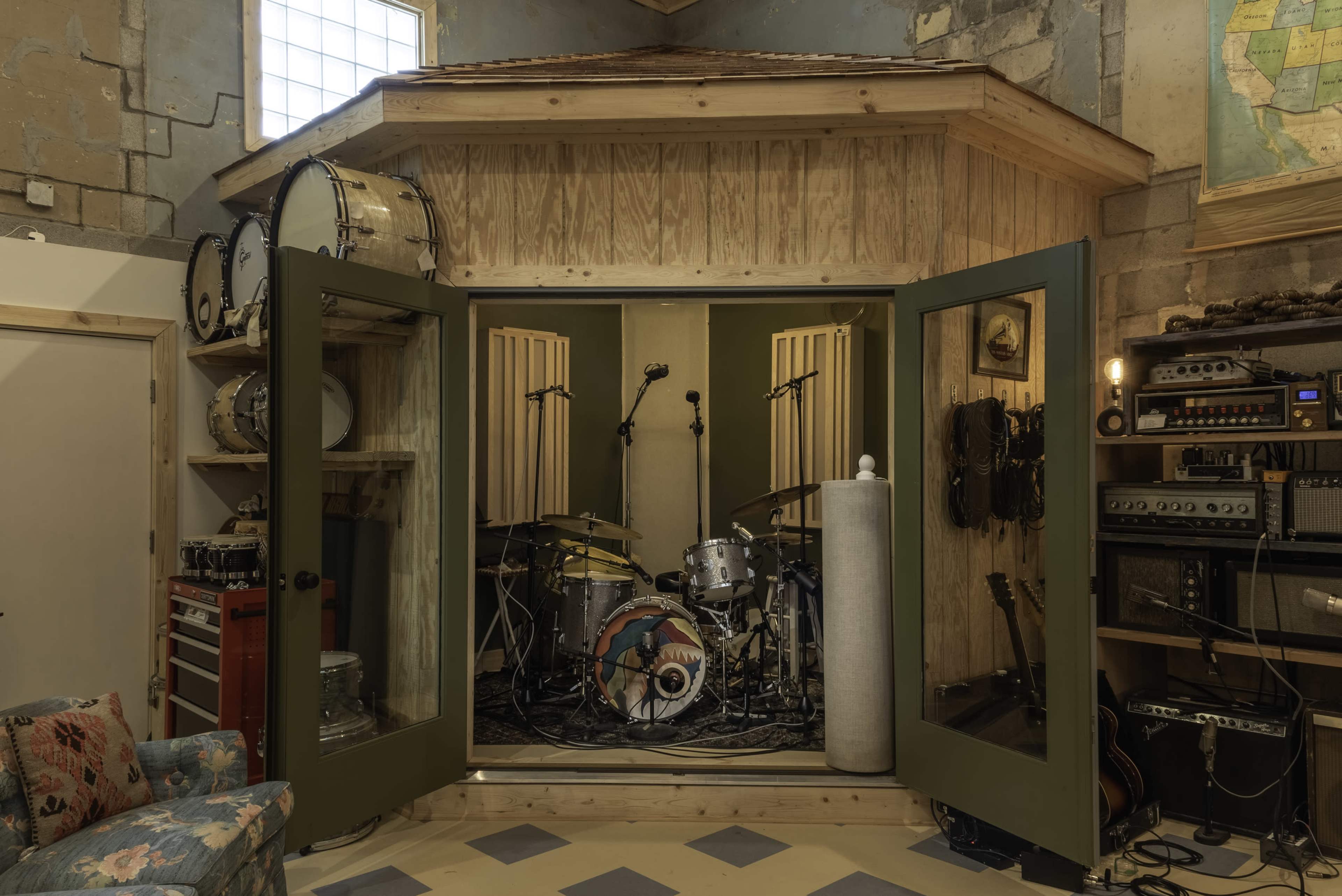 The image shows a soundproof recording studio featuring a drum set enclosed by sliding green doors and various audio equipment displayed on a nearby wall.