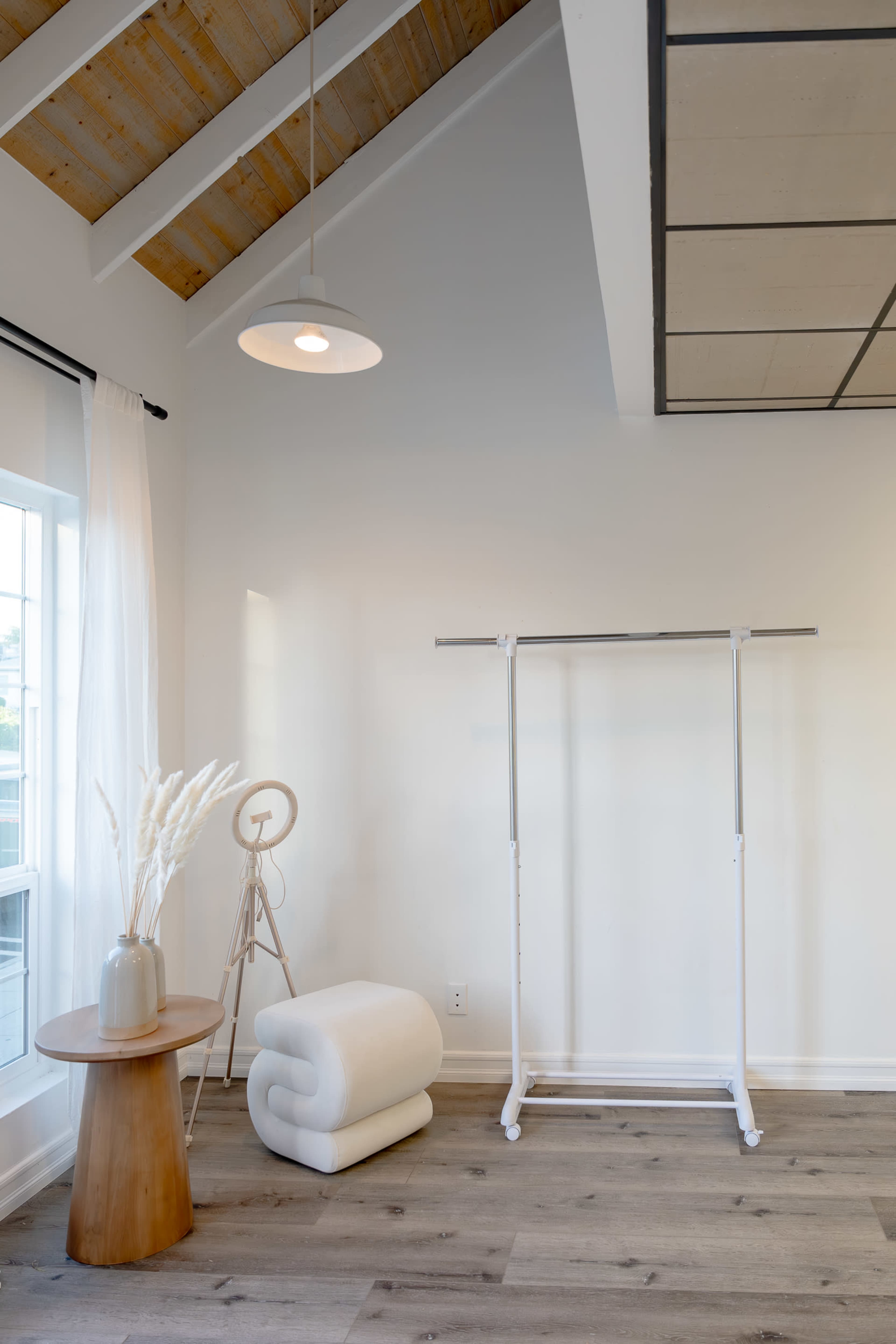 The image shows a minimalist interior featuring a wooden table, a clothing rack, a folded blanket, and a floor lamp with a white shade.