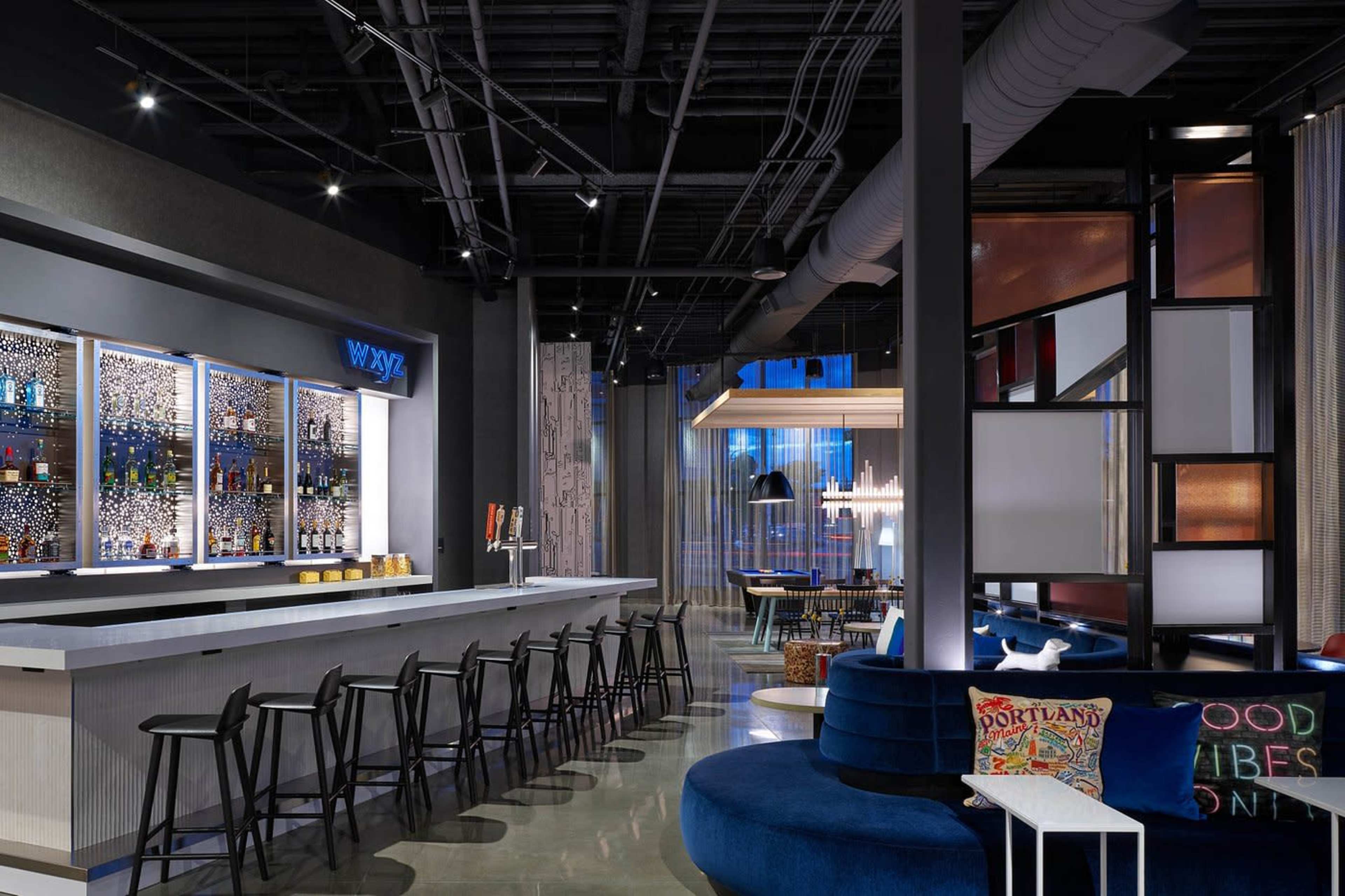 A modern bar area features a well-lit counter with high stools, a vibrant display of bottles, and contemporary seating arrangements.