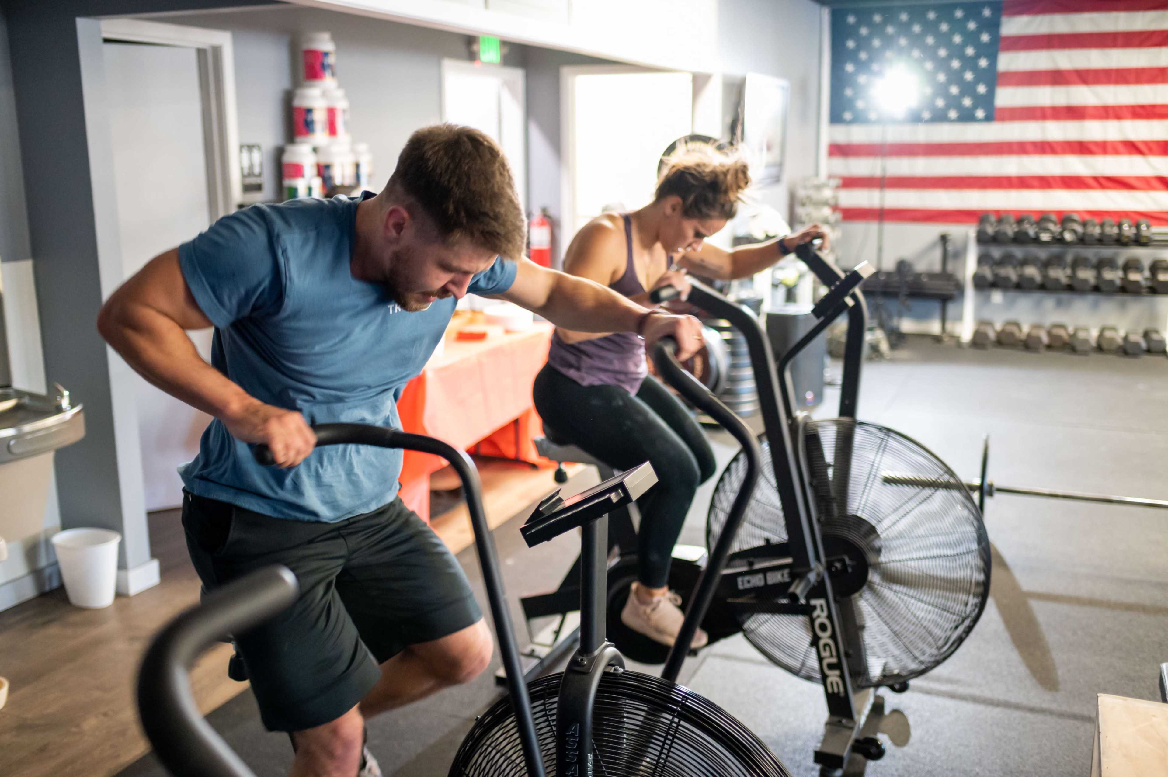 Two individuals are exercising on air bikes in a gym, with fitness equipment and an American flag visible in the background.