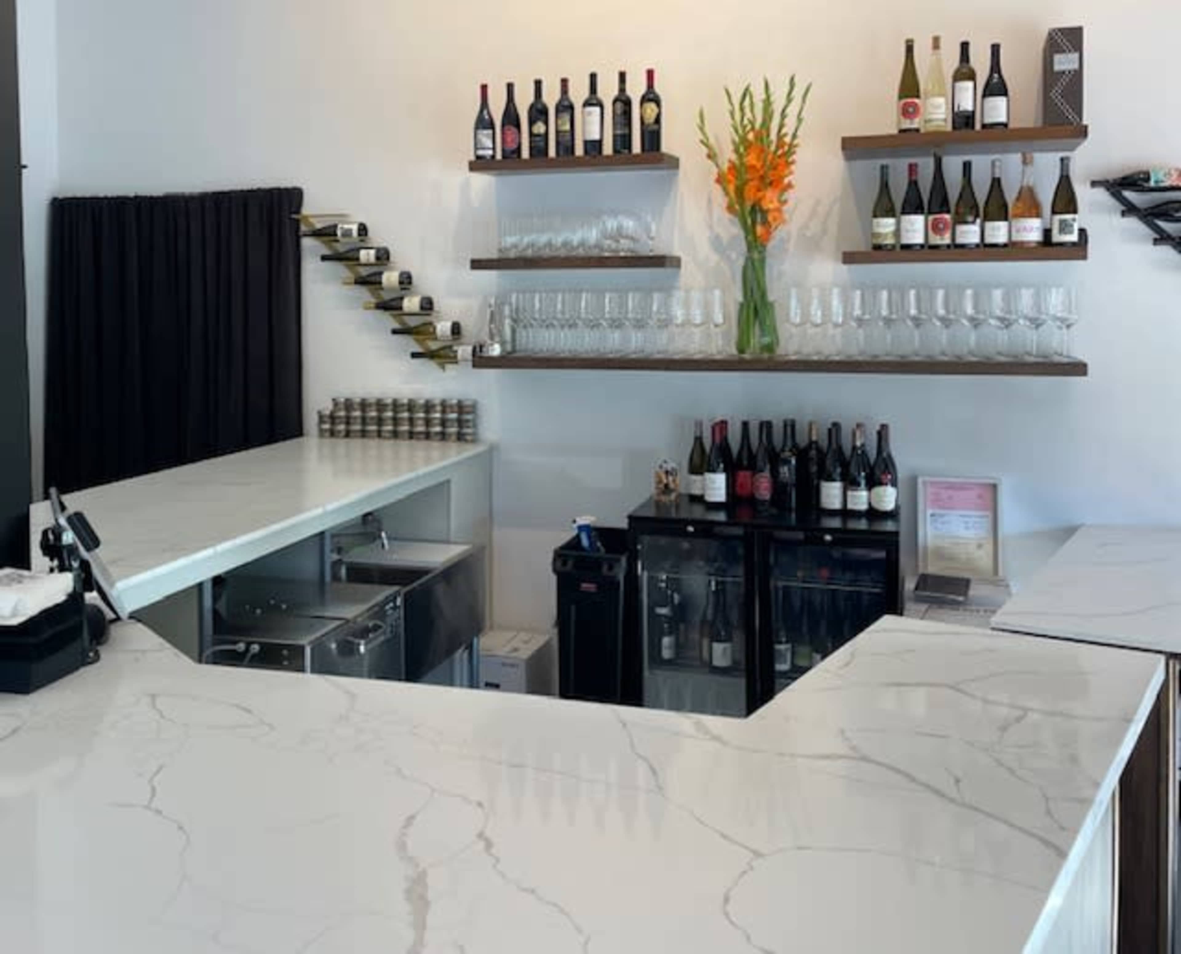 The image shows a modern bar area with a marble countertop, shelves displaying various wine bottles, and a cooler holding additional bottles, accented by a vase of orange flowers.