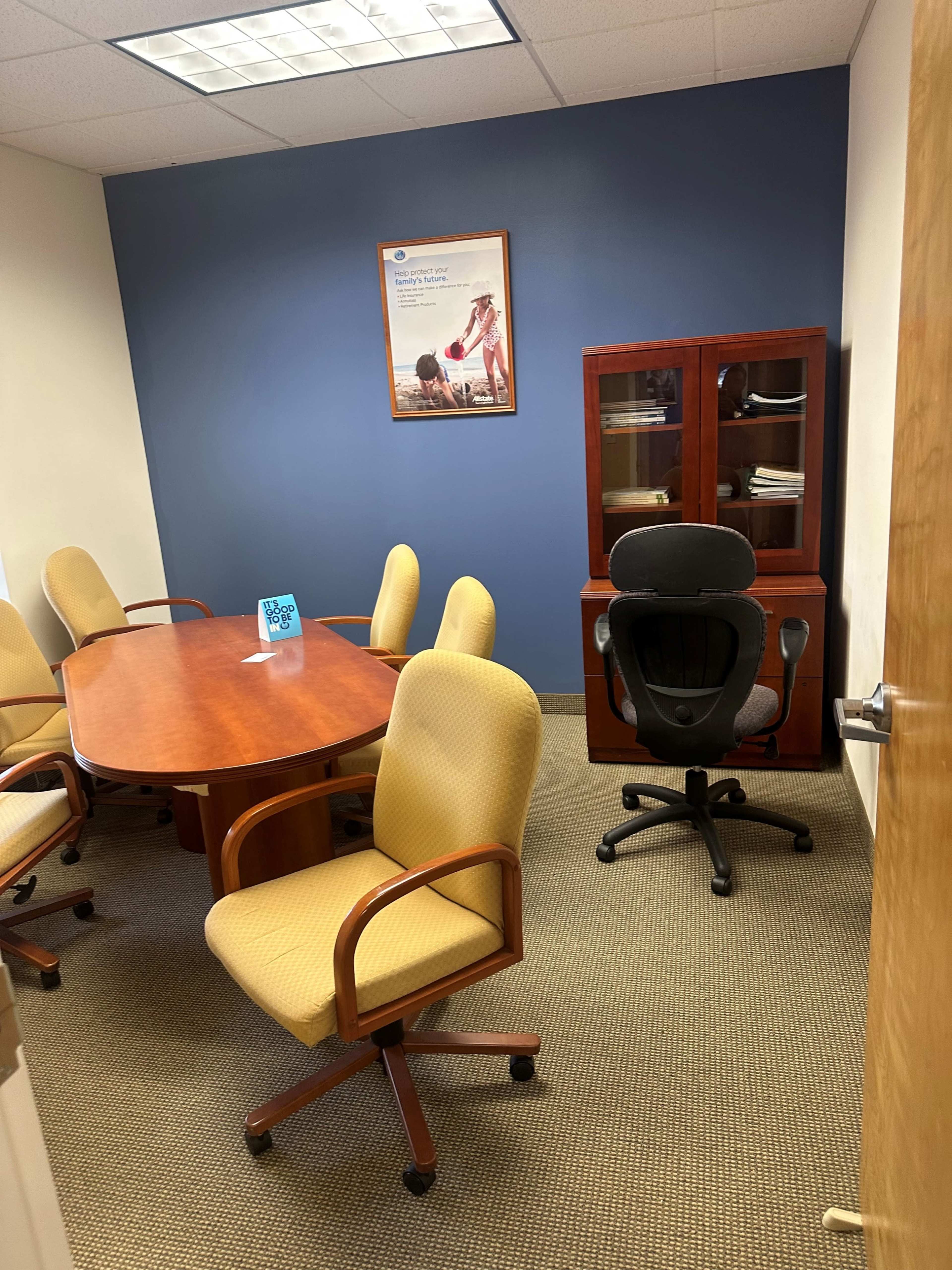 The image shows a small conference room with a wooden table surrounded by yellow upholstered chairs and a dark wooden cabinet against a blue wall.