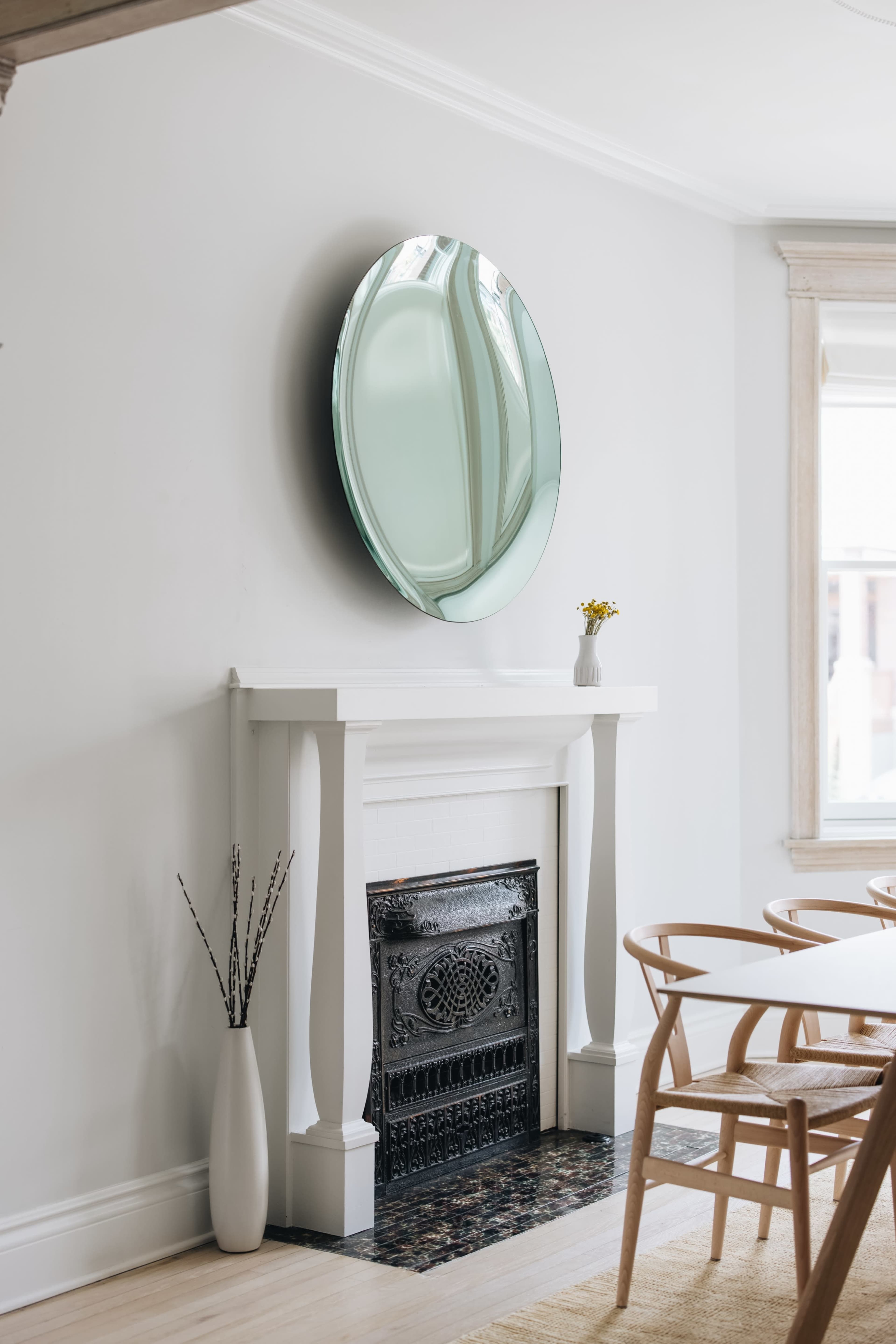 A round mirror hangs above a white fireplace, flanked by a decorative vase and a simple floral arrangement on a dining table.