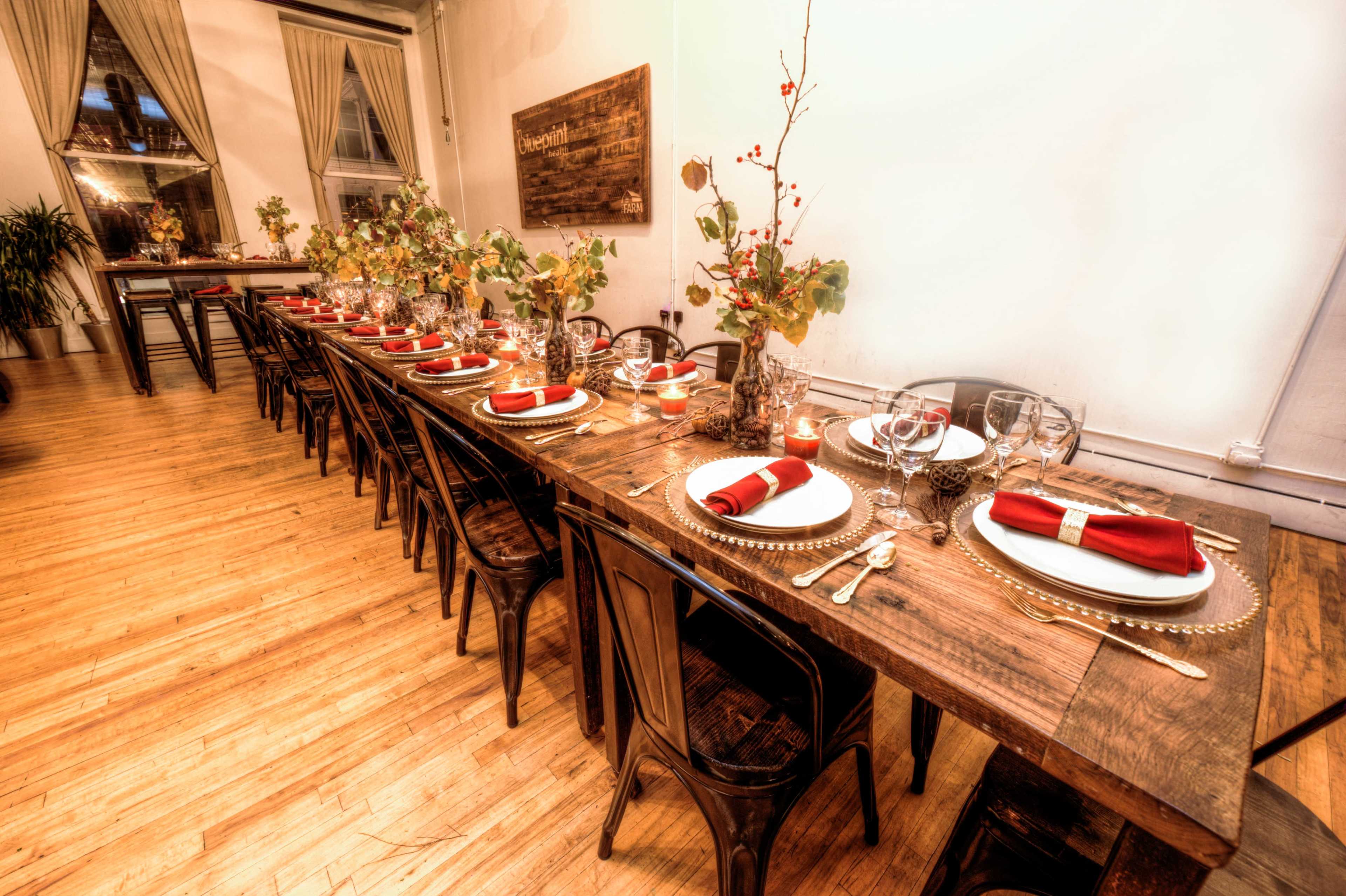 A long wooden dining table is elegantly set with red napkins, glassware, and decorative foliage arrangements.