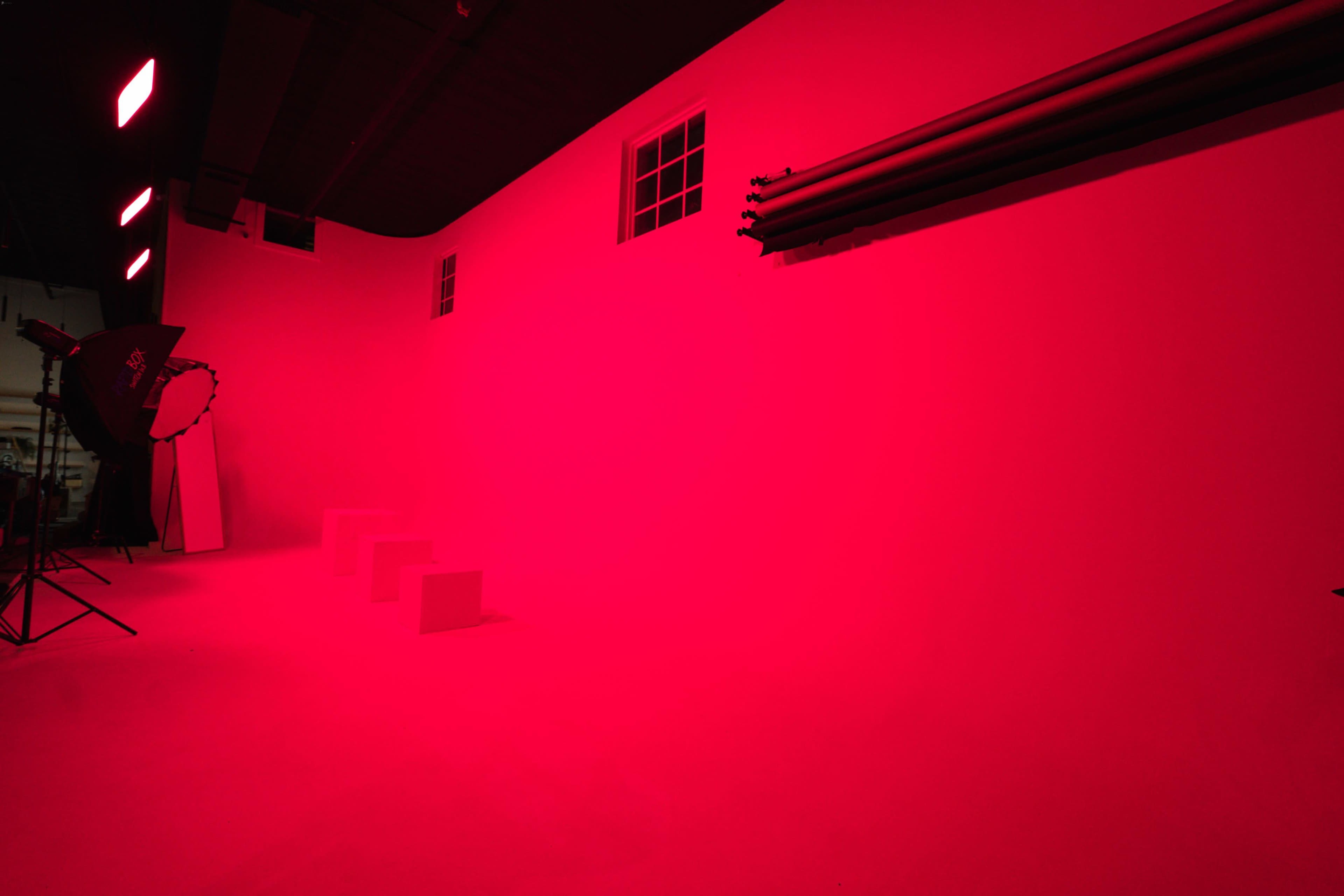 A photography studio is illuminated with red lighting, featuring a blank wall, steps, and equipment like light stands and reflector.