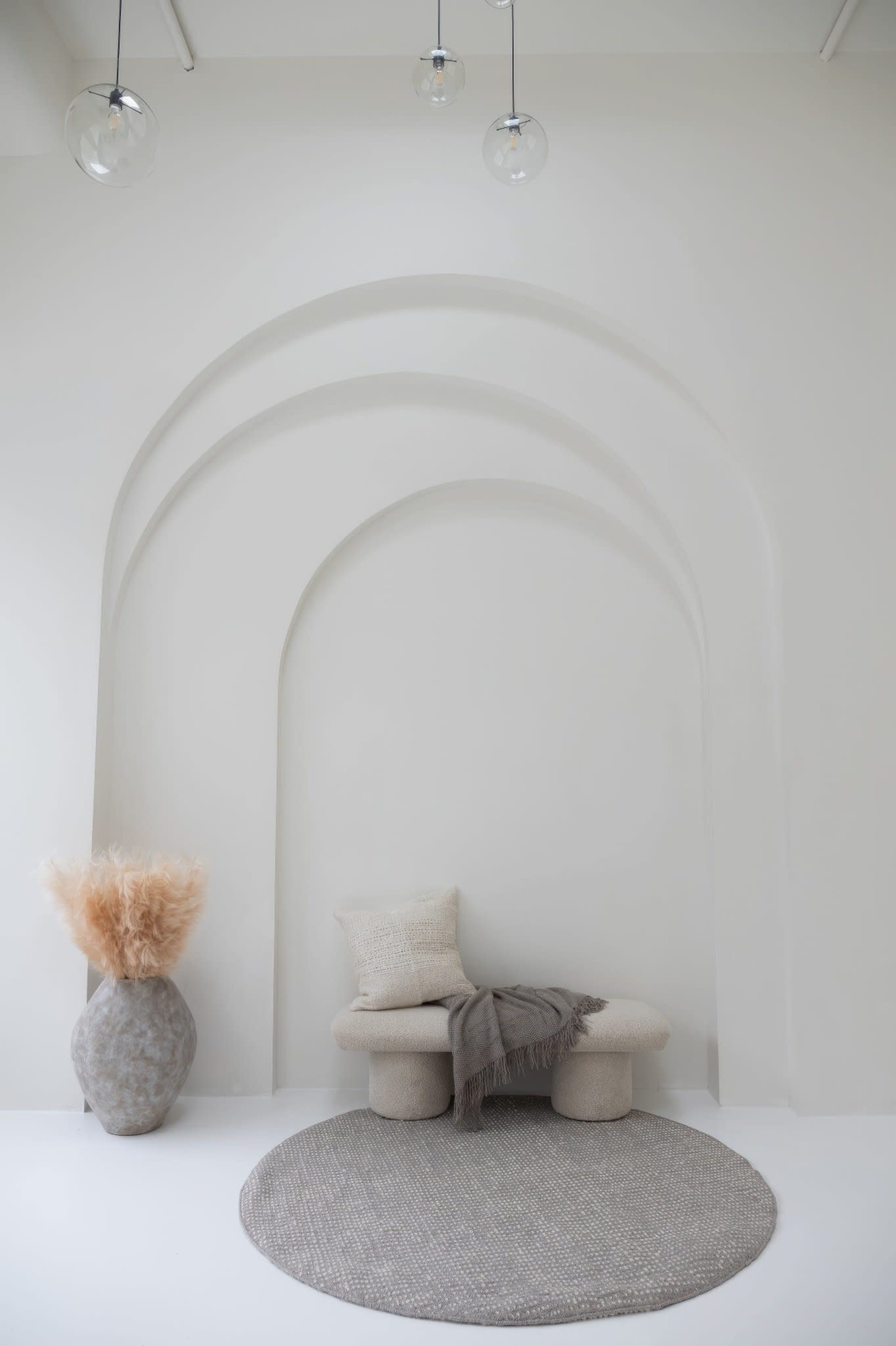 A minimalist interior featuring a curved alcove, a sculptural seating area, a decorative vase with pampas grass, and a circular rug on a light-colored floor.