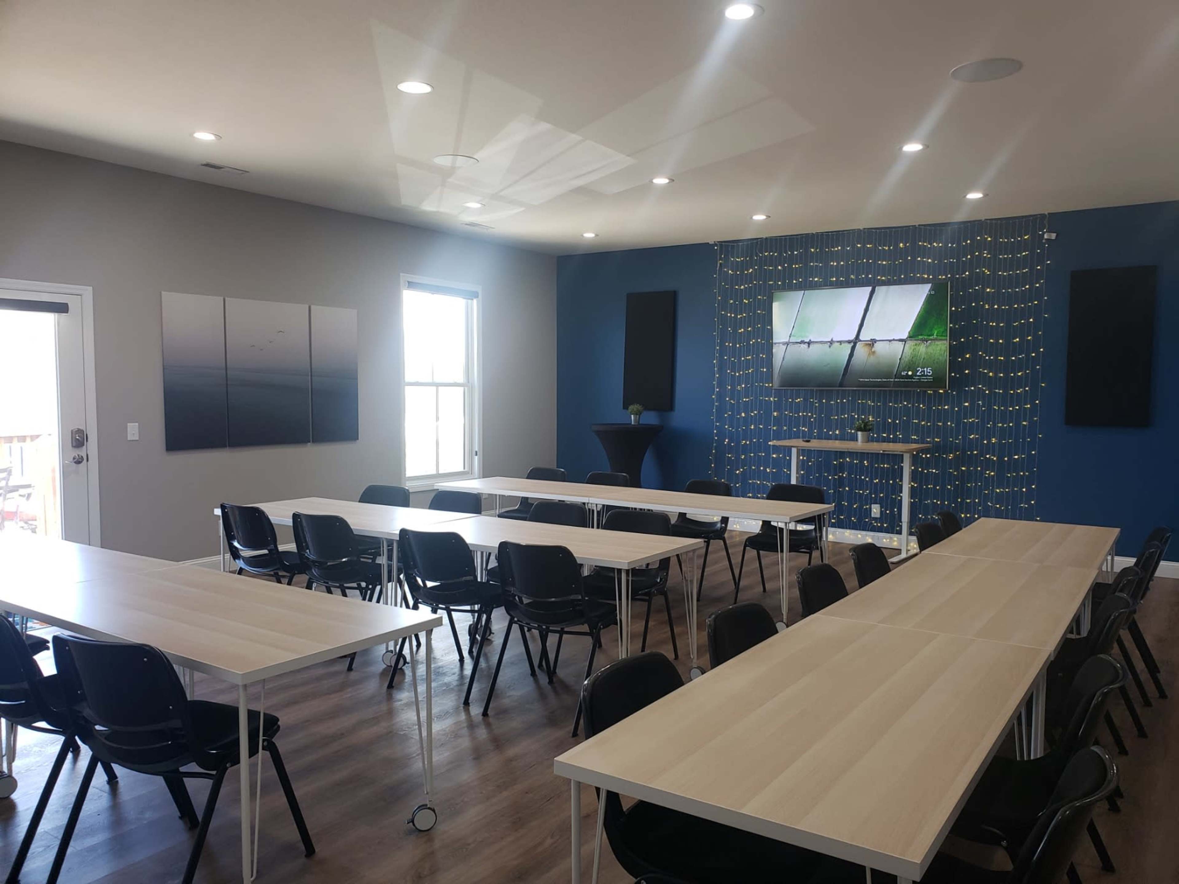 The image shows a modern meeting room with multiple tables arranged in a U-shape, black chairs, and a large screen mounted on a blue accent wall adorned with decorative lighting.