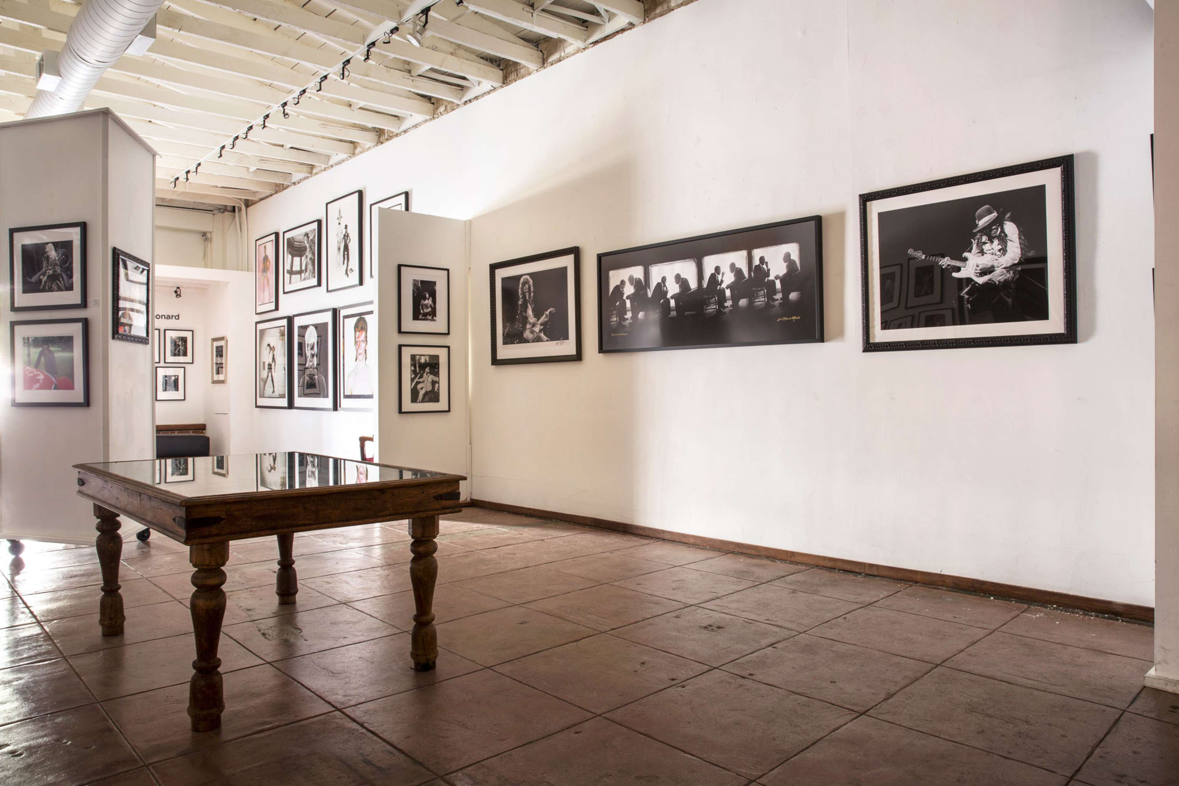 An art gallery featuring black-and-white photographs displayed on white walls, with a wooden table in the center.