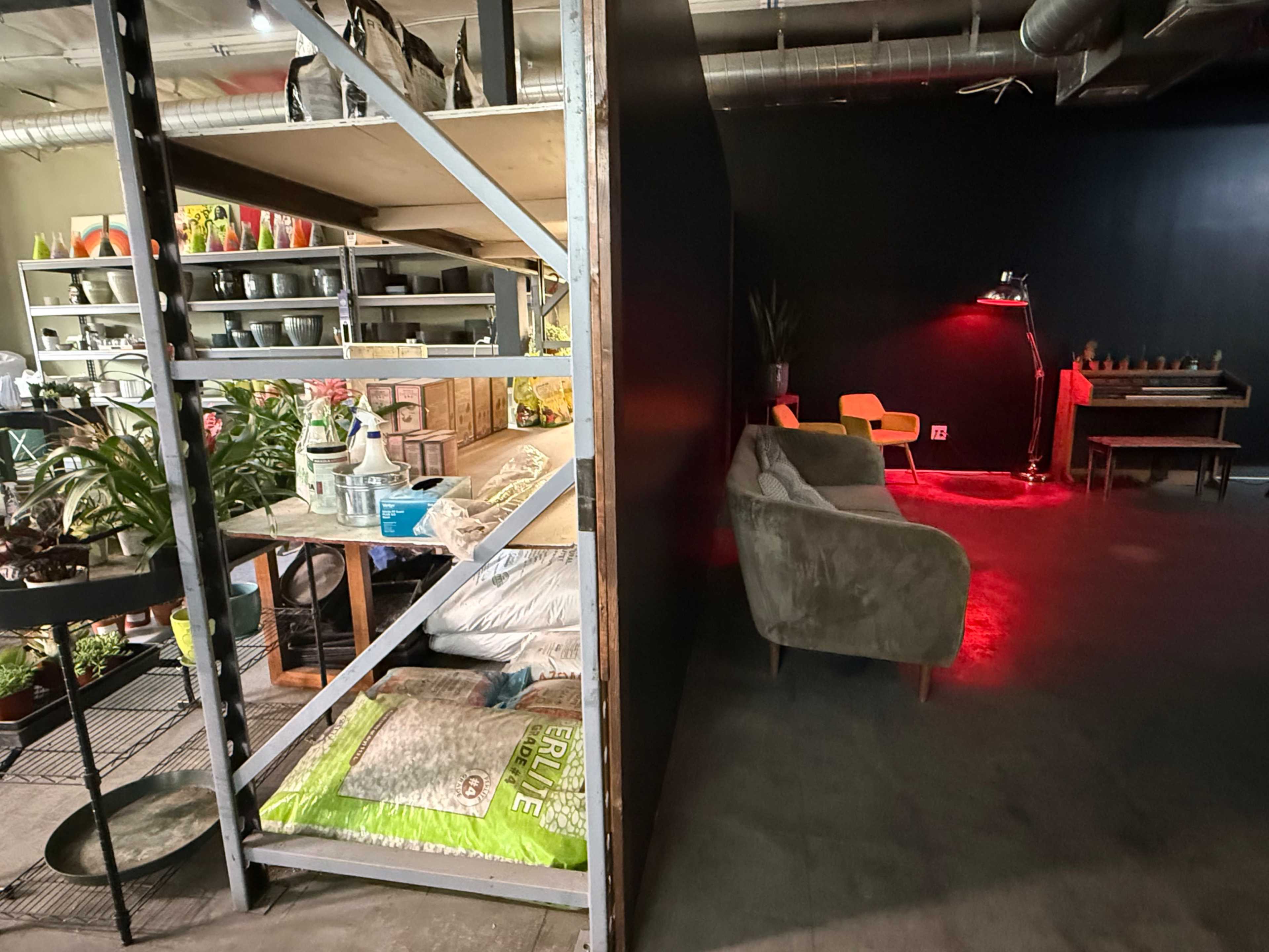 The image shows a divided interior space with shelving units stocked with potted plants and gardening supplies on one side, and a cozy seating area illuminated by a red lamp on the other.