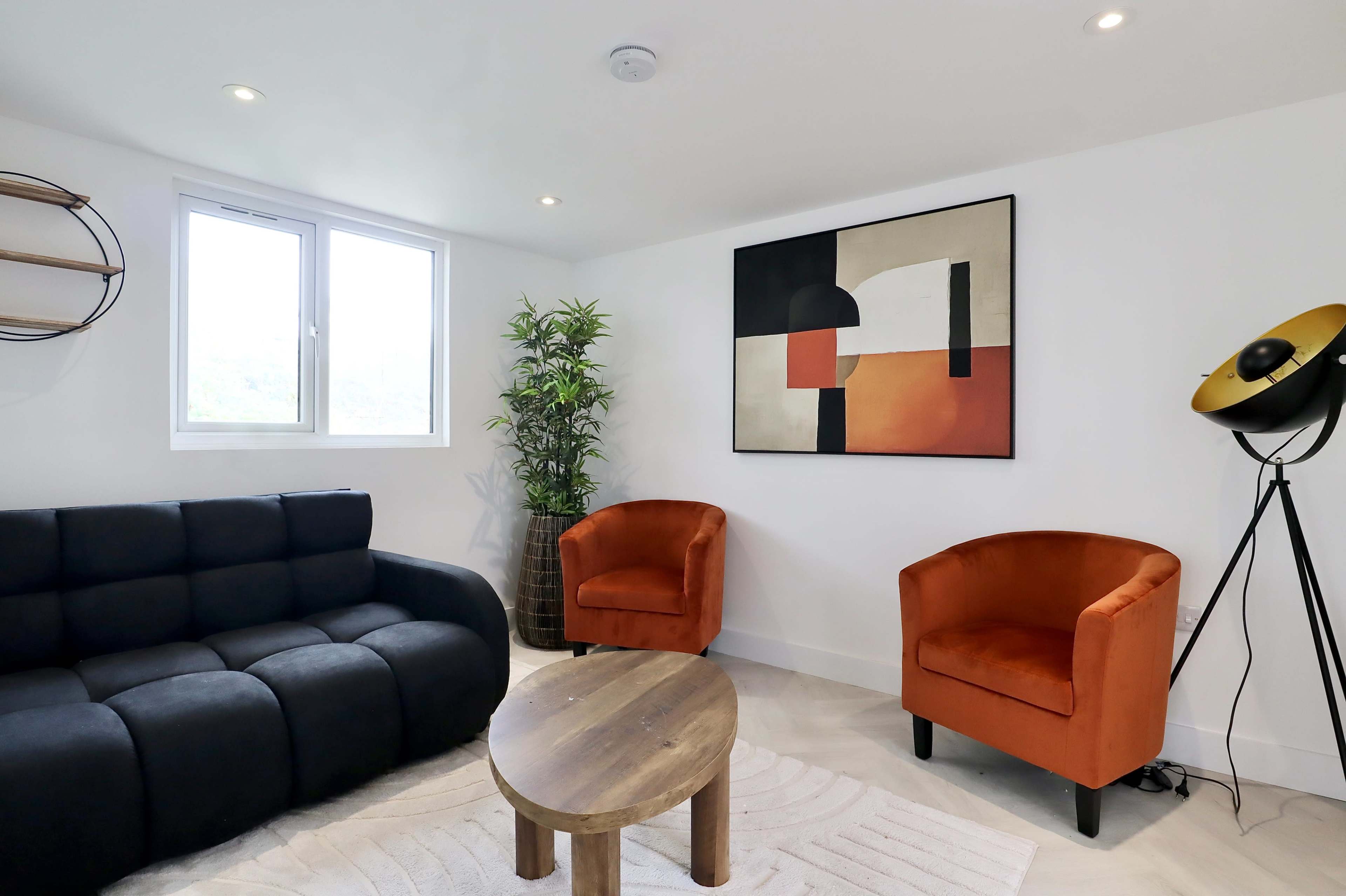 The image shows a modern living room featuring a black sofa, two orange armchairs, a round wooden coffee table, and abstract wall art, with a large window allowing natural light to enter.