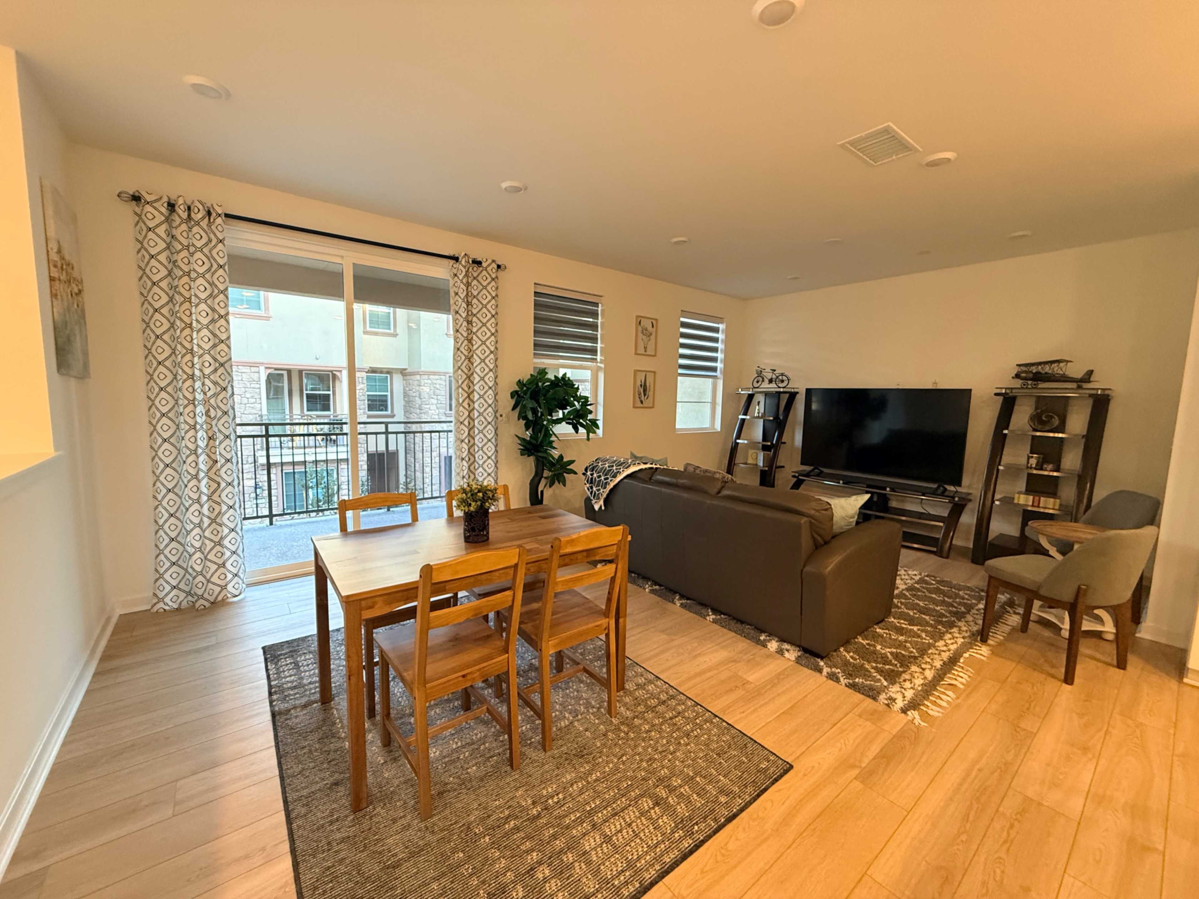 The image shows a modern living space with a dining table and chairs, a couch facing a television, and large windows leading to a balcony.