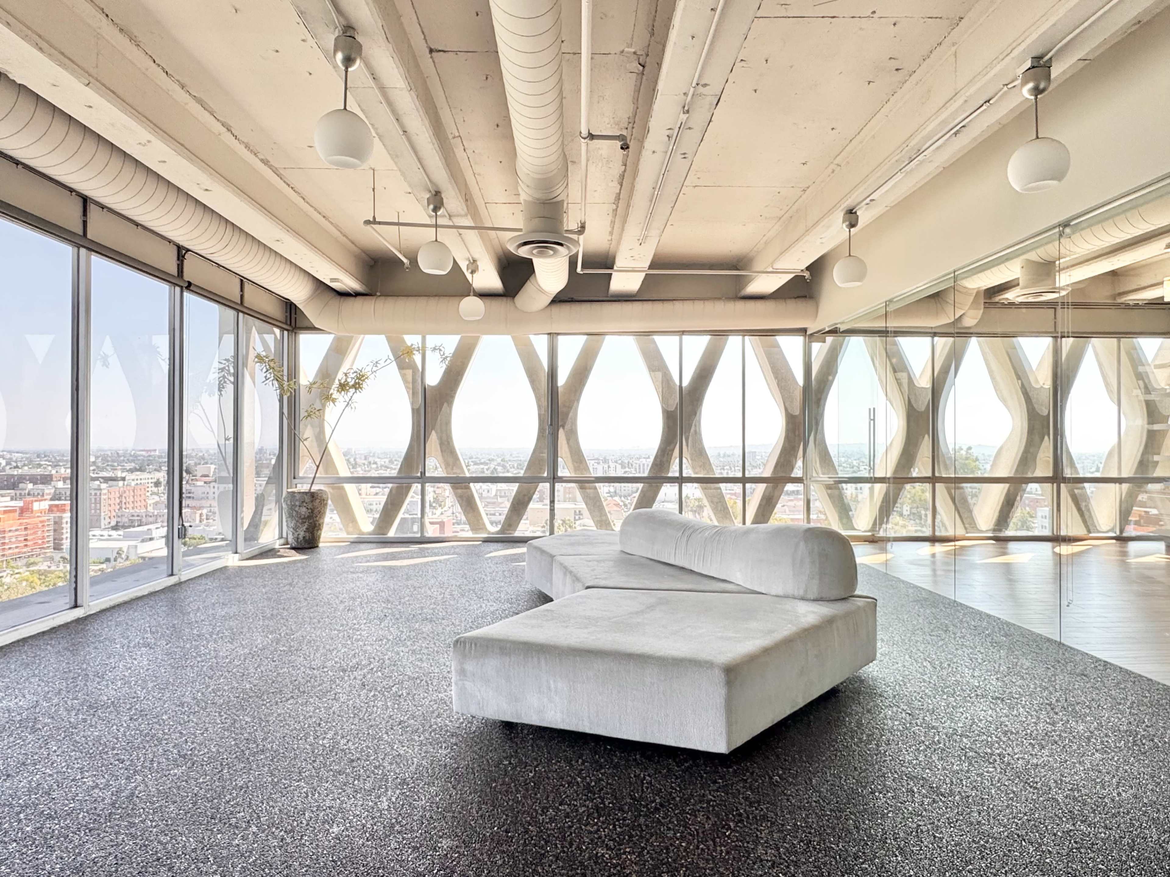 The image shows a spacious, modern interior with a large, curved white sofa and floor-to-ceiling windows offering a view of the city.