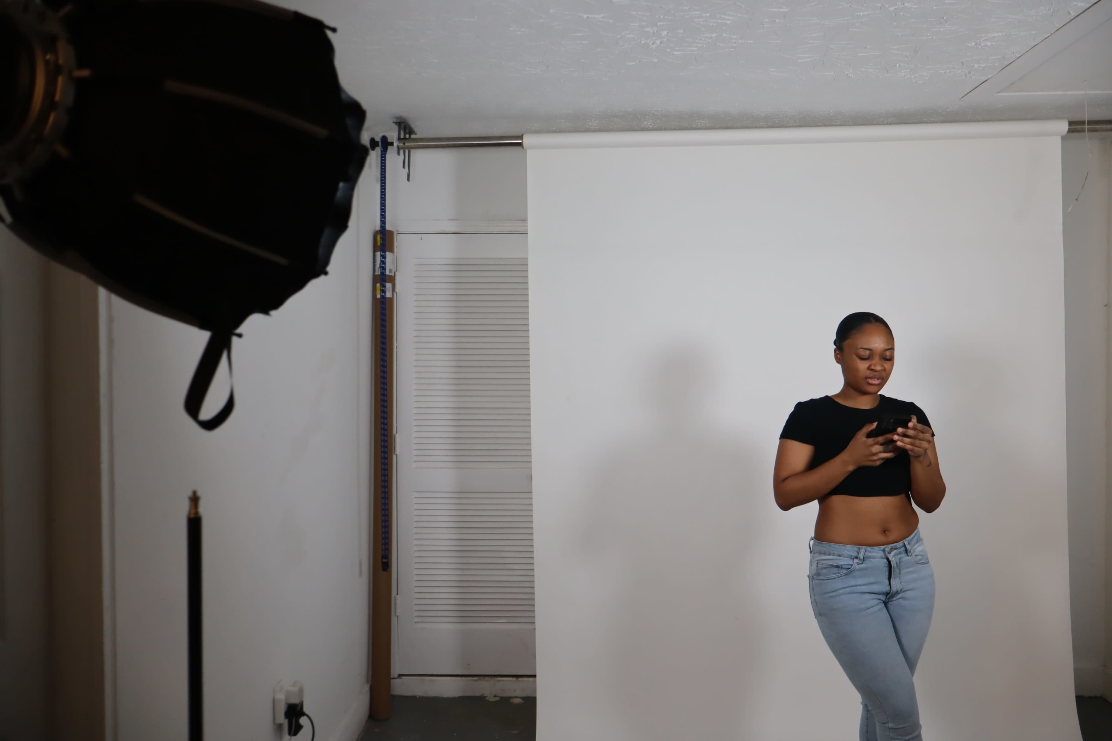 A woman in a black crop top and blue jeans stands in a photography studio, looking at her phone against a plain white backdrop.