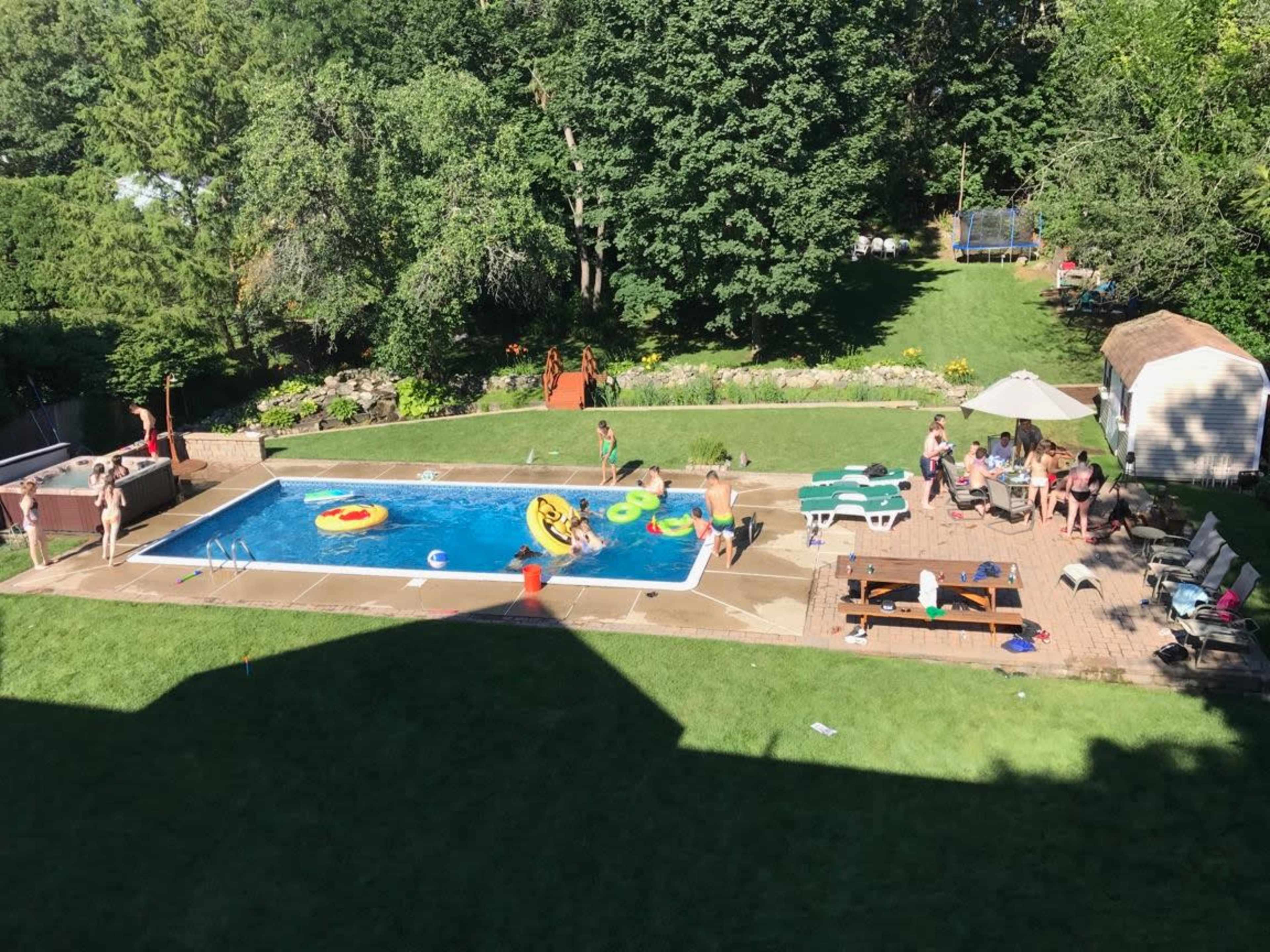 A group of people enjoy a sunny day by a swimming pool surrounded by greenery, with various floats and lounge chairs arranged around the area.