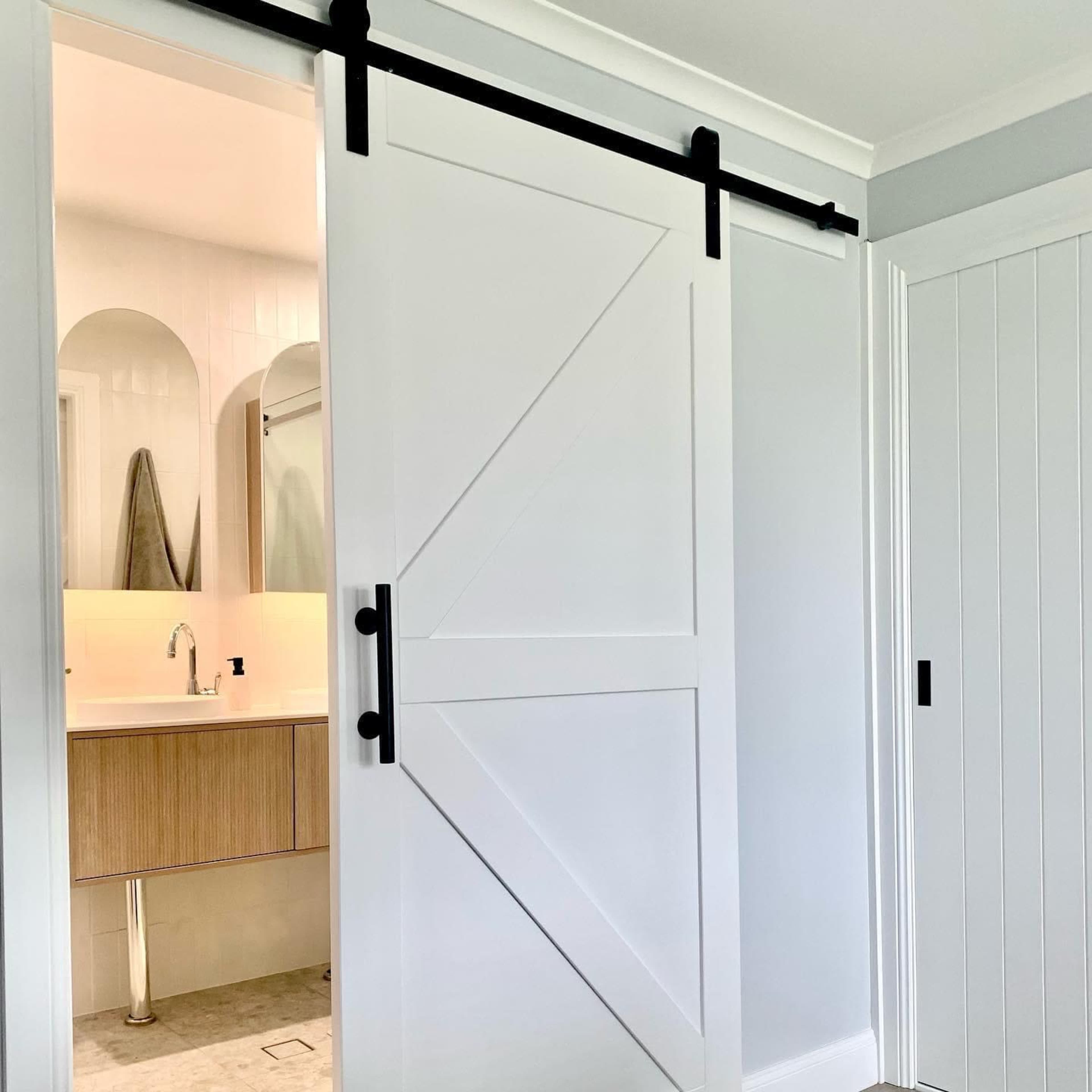 A white barn door with black hardware partially open, revealing a bathroom with a wooden vanity and two arched mirrors.