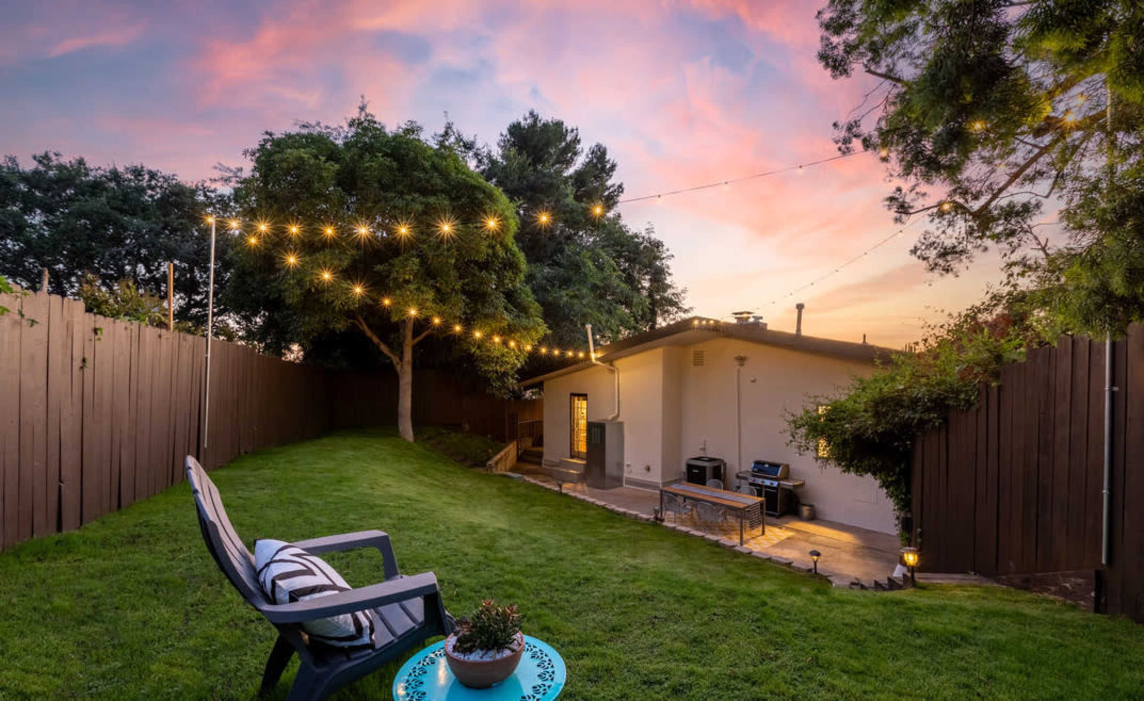 A well-maintained backyard features a grassy area, a patio with a grill, and string lights suspended between trees at sunset.
