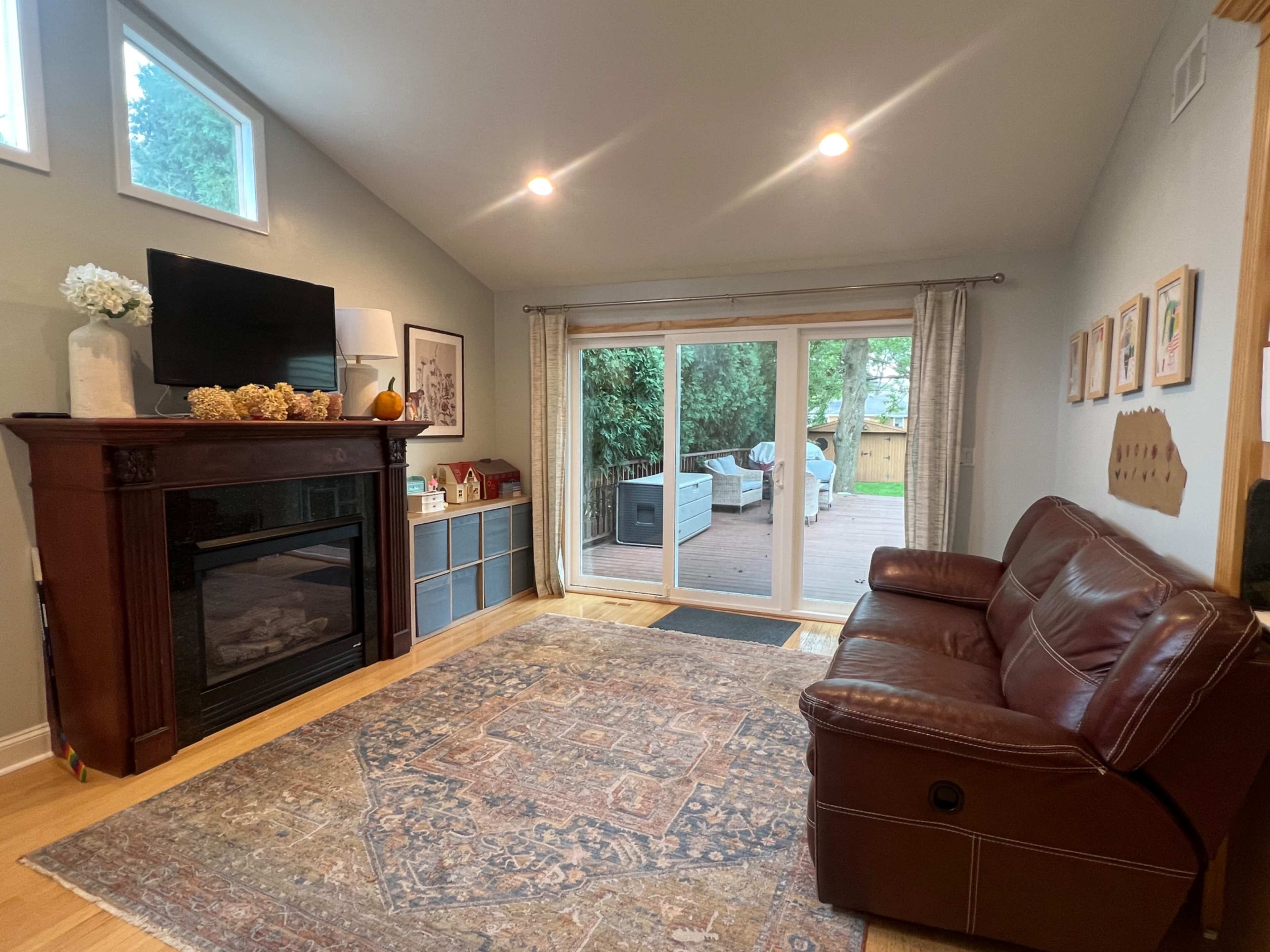 A cozy living room features a brown leather couch, a fireplace with decorations, and sliding glass doors that open to a wooden deck outside.