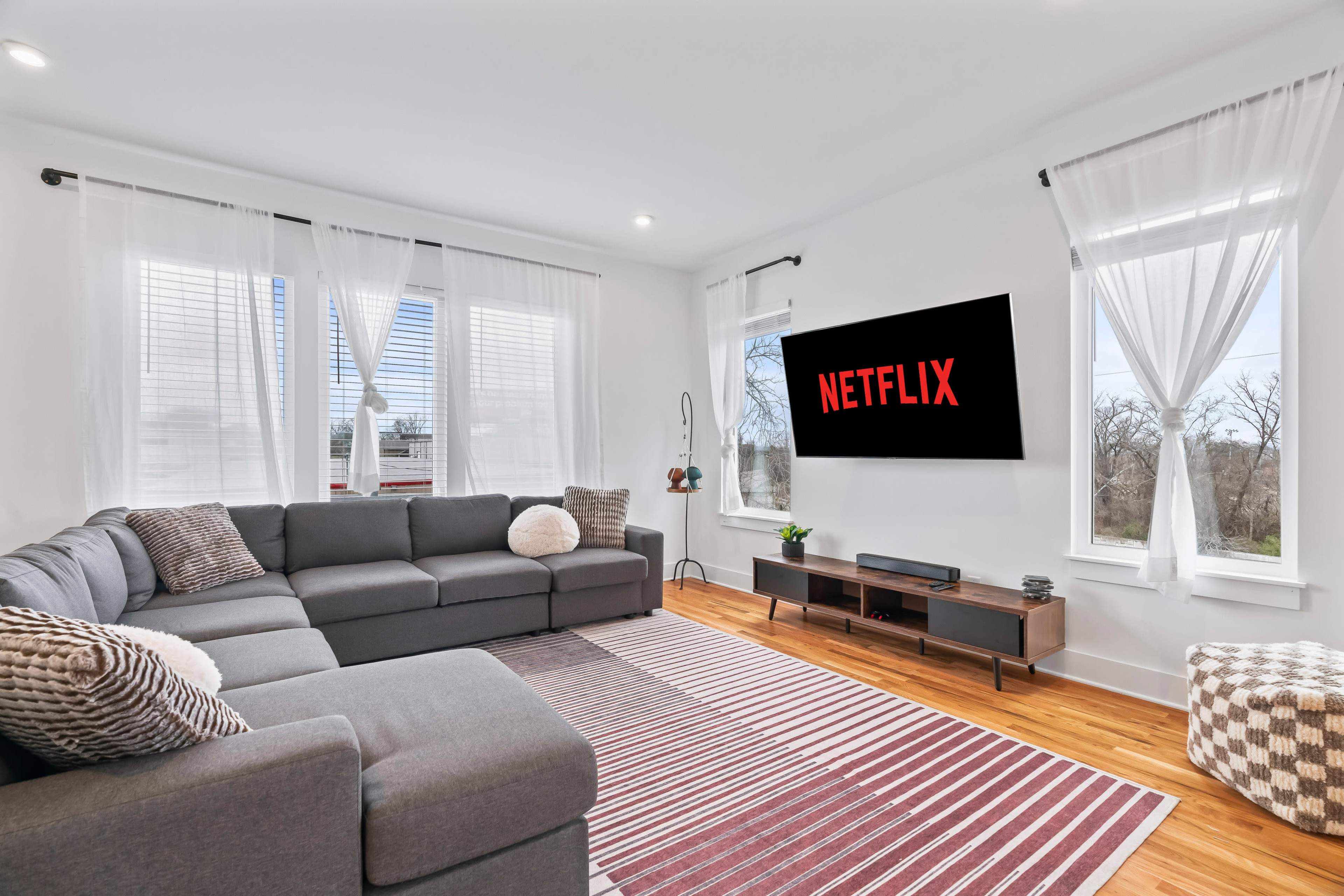 A modern living room features a large sectional sofa, a wooden TV stand, and a wall-mounted television displaying the Netflix logo, with large windows dressed in sheer curtains.