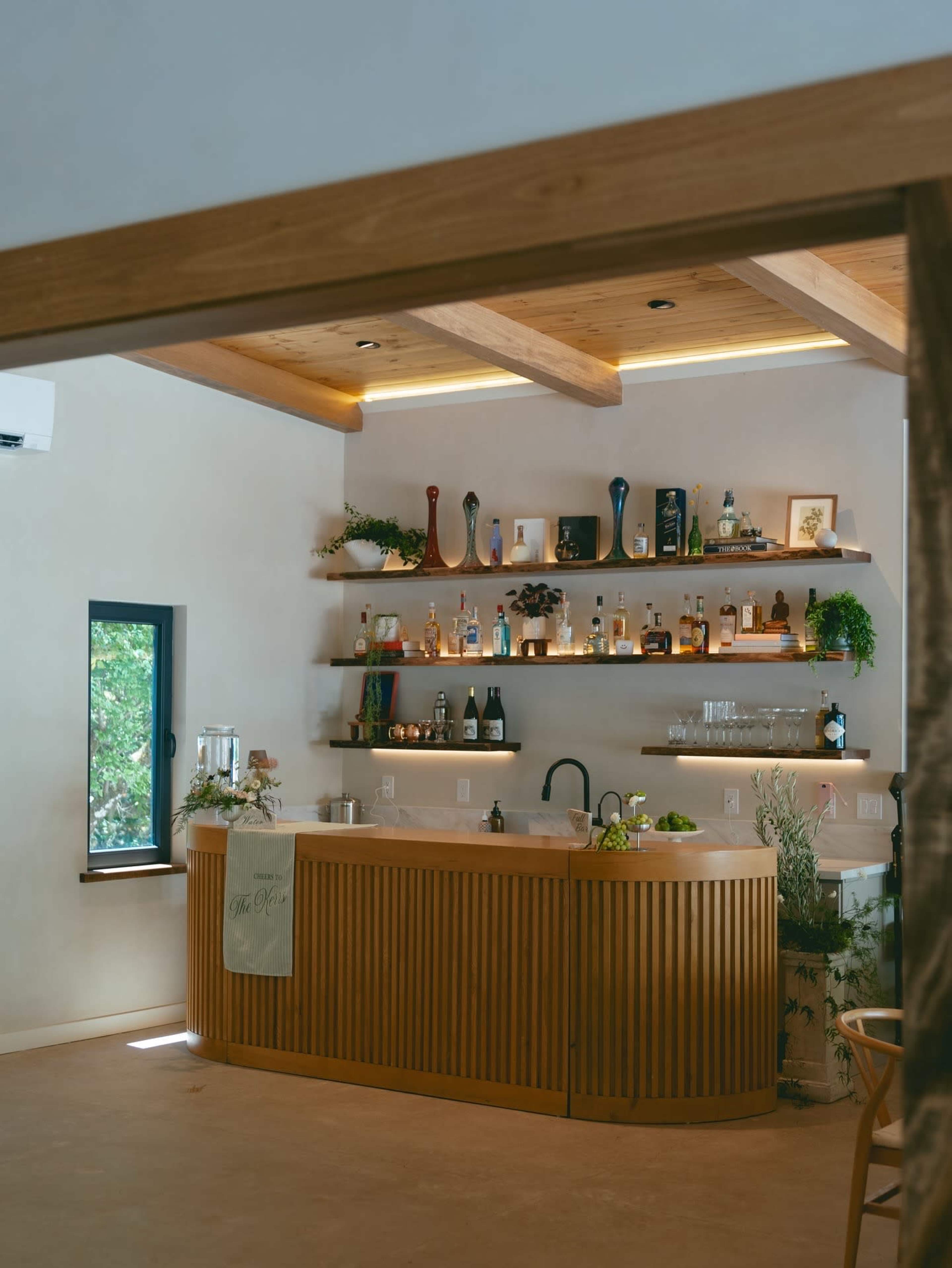 The image shows a wooden bar with various bottles displayed on shelves above it, set in a well-lit interior with greenery around.