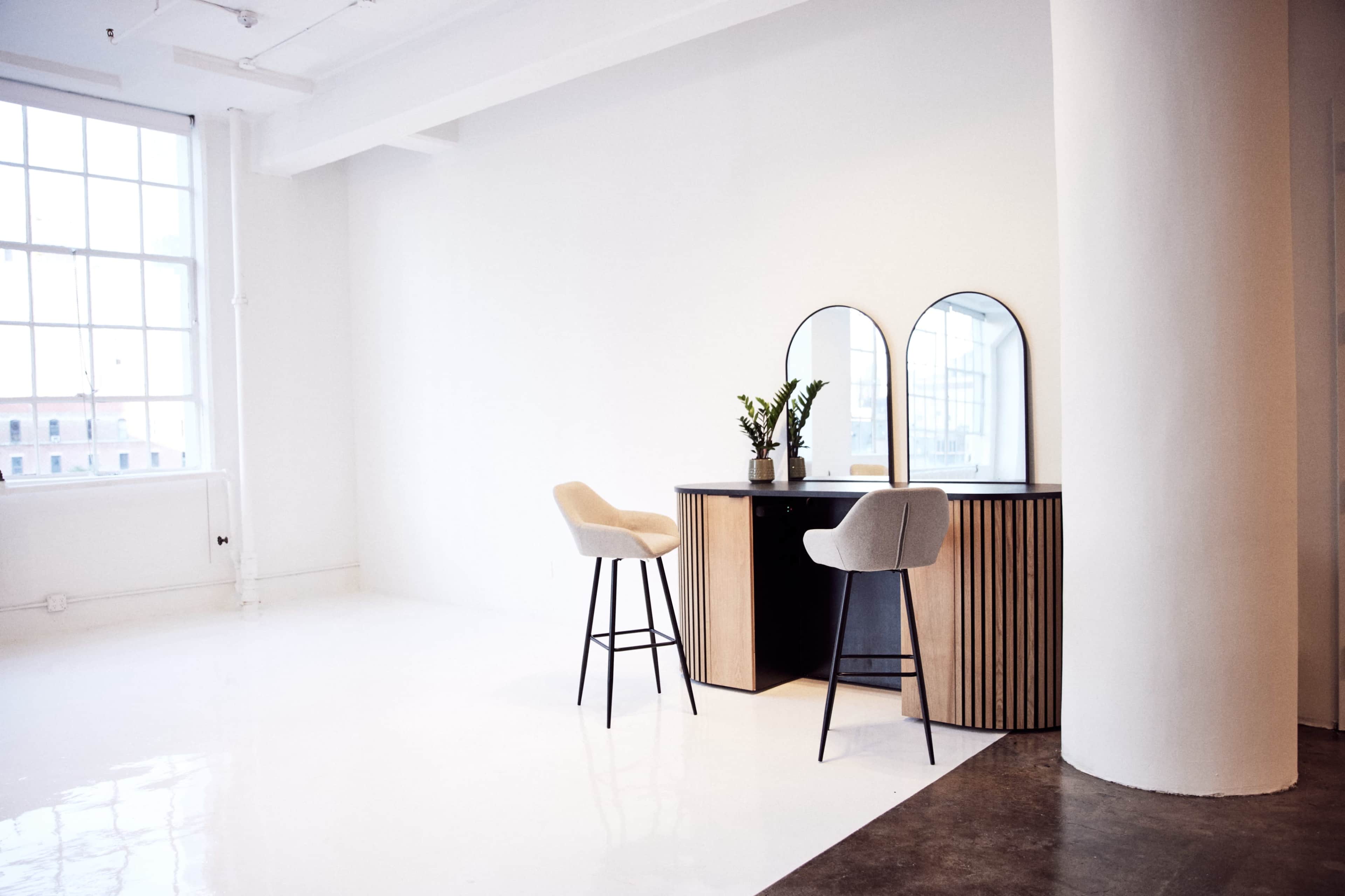 A minimalist interior features a wooden vanity with two round mirrors and two stools against a white wall, illuminated by natural light from a large window.