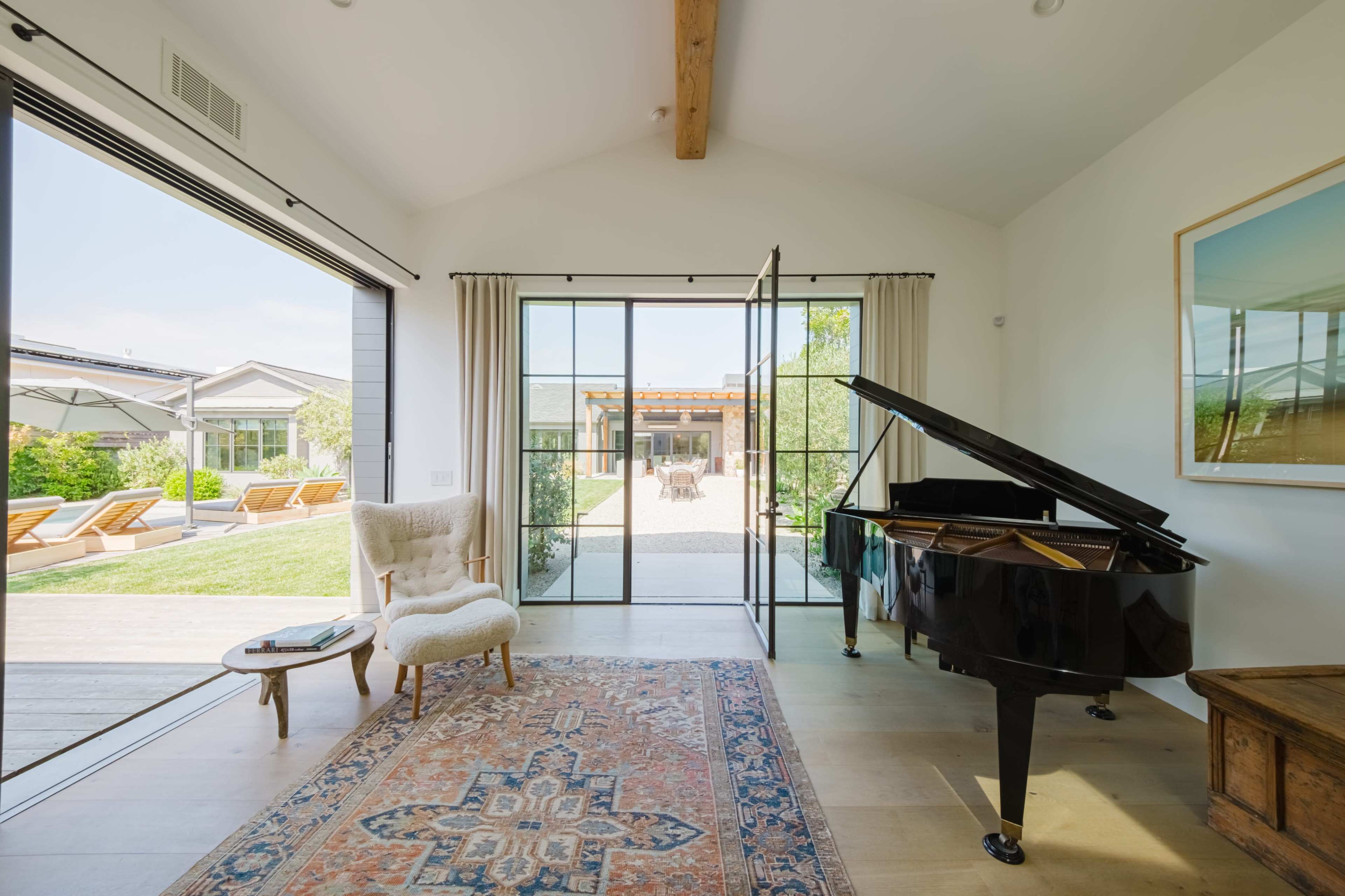 A grand piano sits beside a chair and rug in a brightly lit room with large sliding glass doors opening to a manicured outdoor area.