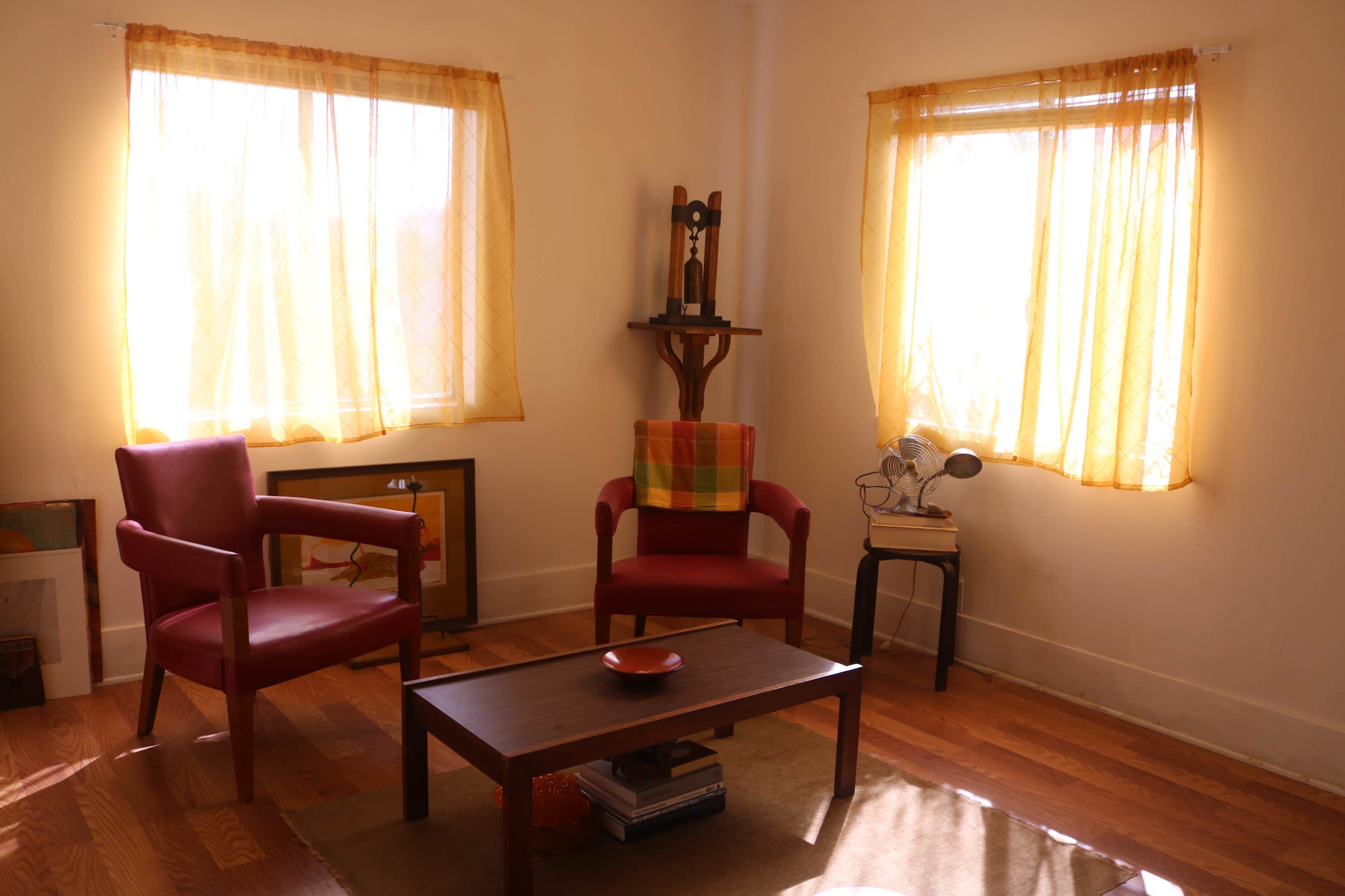 A cozy living room with two red chairs, a wooden table, a small bookshelf, and bright yellow curtains framing the windows.