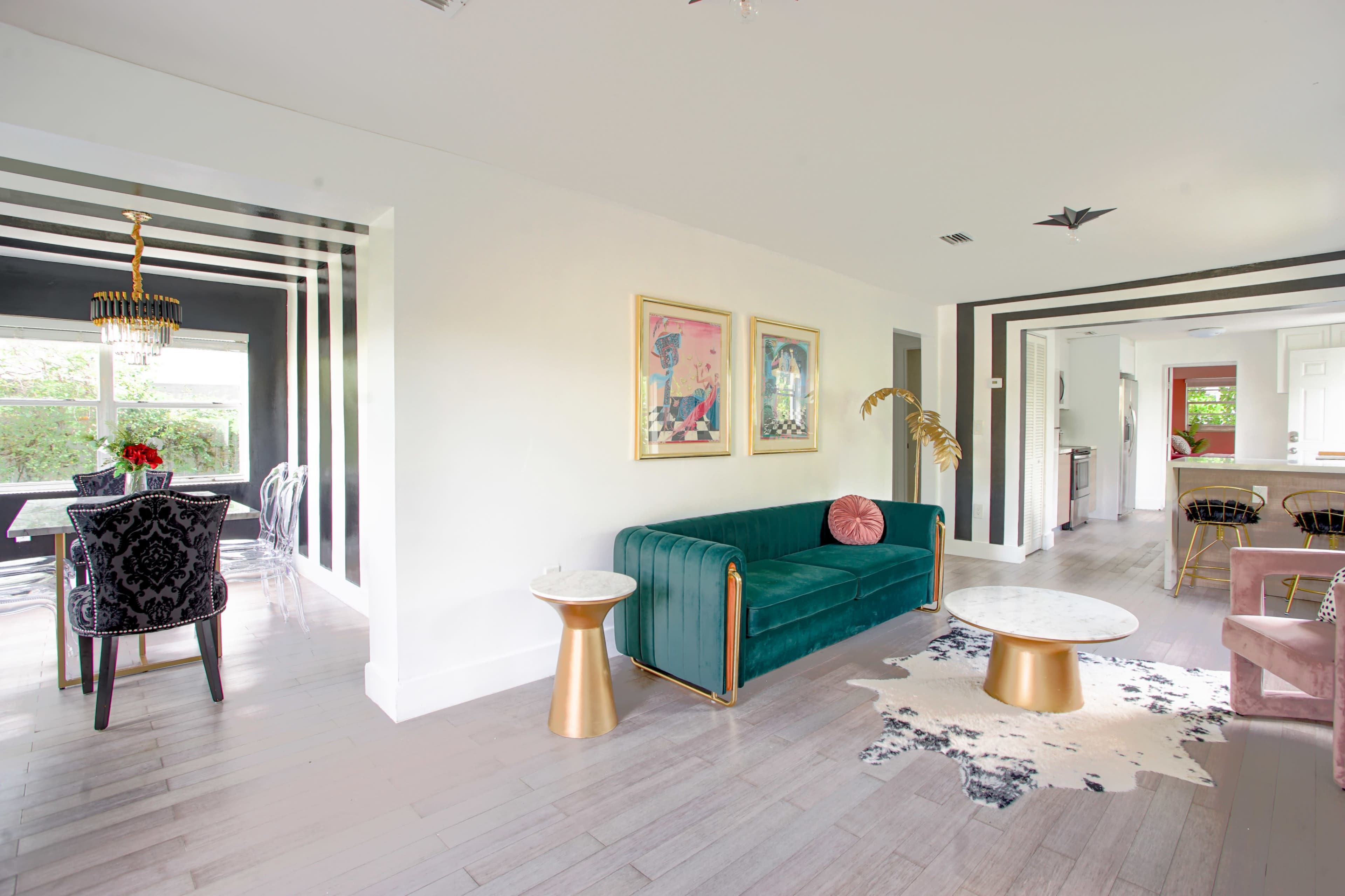 A modern living area featuring a green velvet sofa, marble-topped side tables, and black and white striped walls, leading into a dining area with dark decor.
