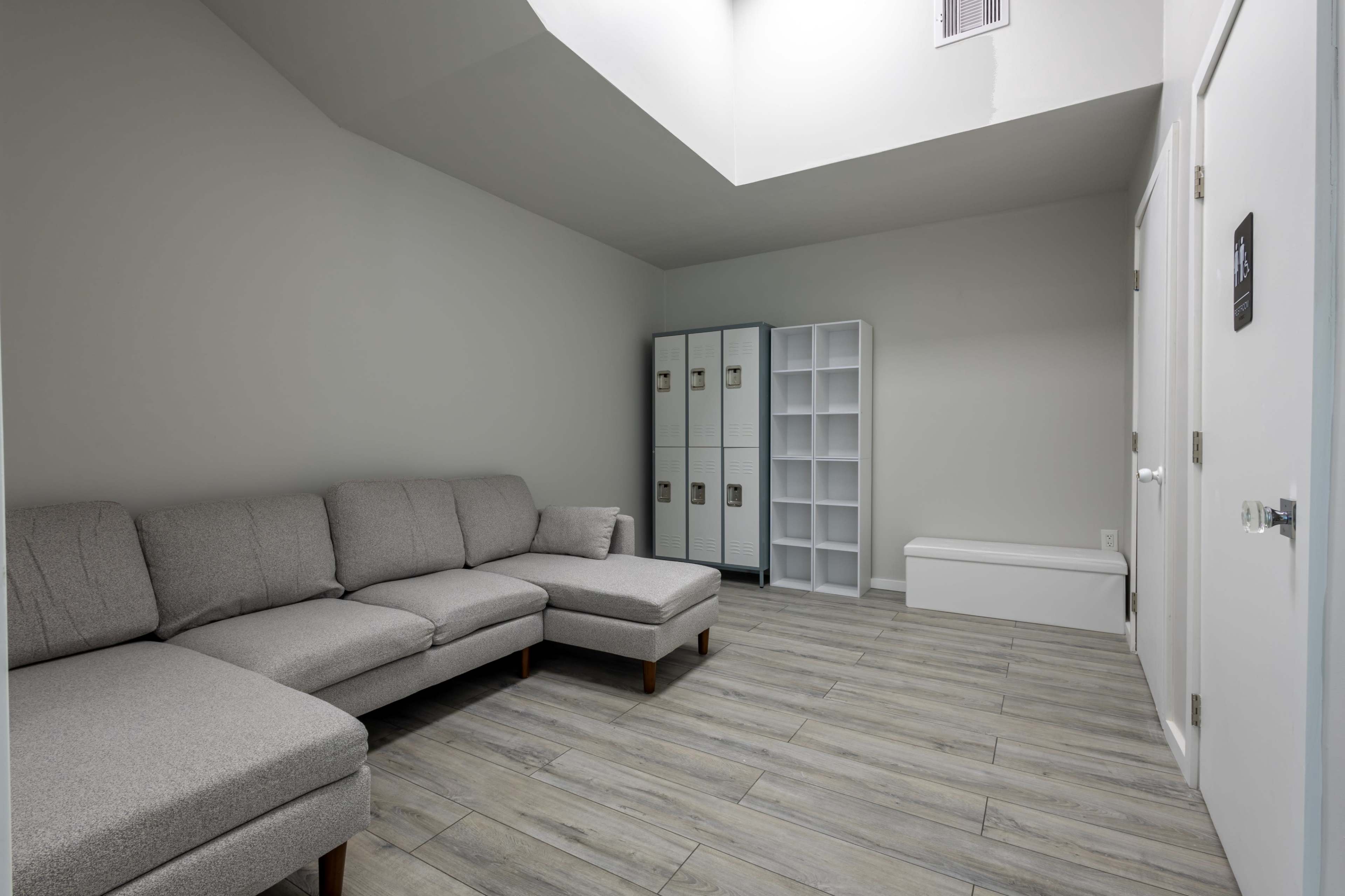 The image shows a simple interior room with a gray sectional sofa, storage lockers, a white shelving unit, and a bench against a wall, all set on a tiled floor.