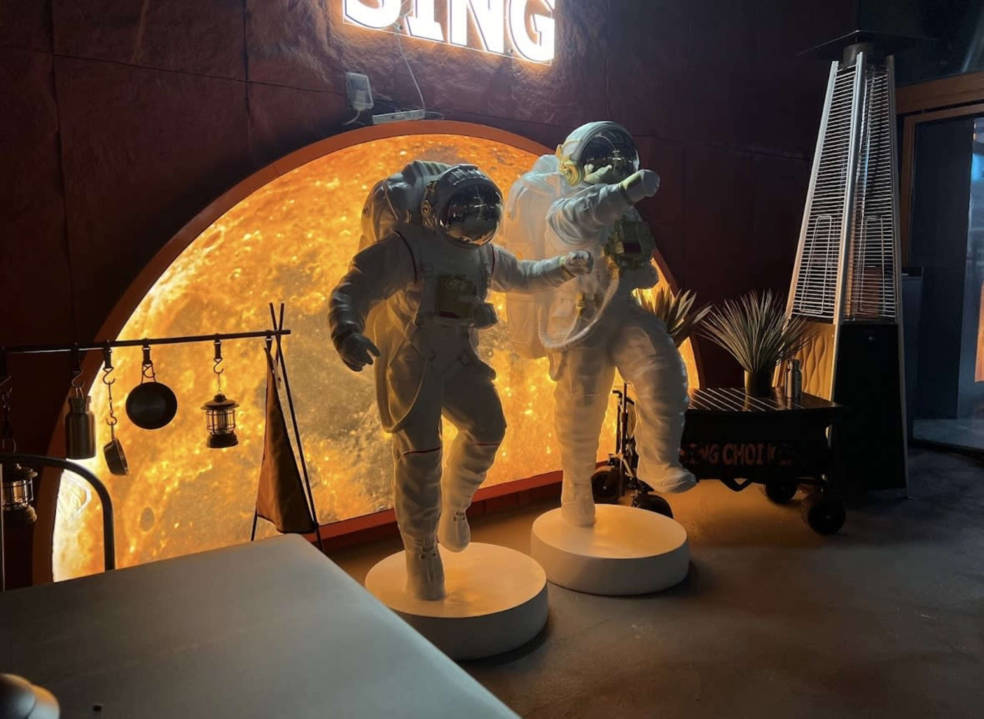Two astronaut figures stand on pedestals in front of a large, illuminated orange backdrop resembling the sun.