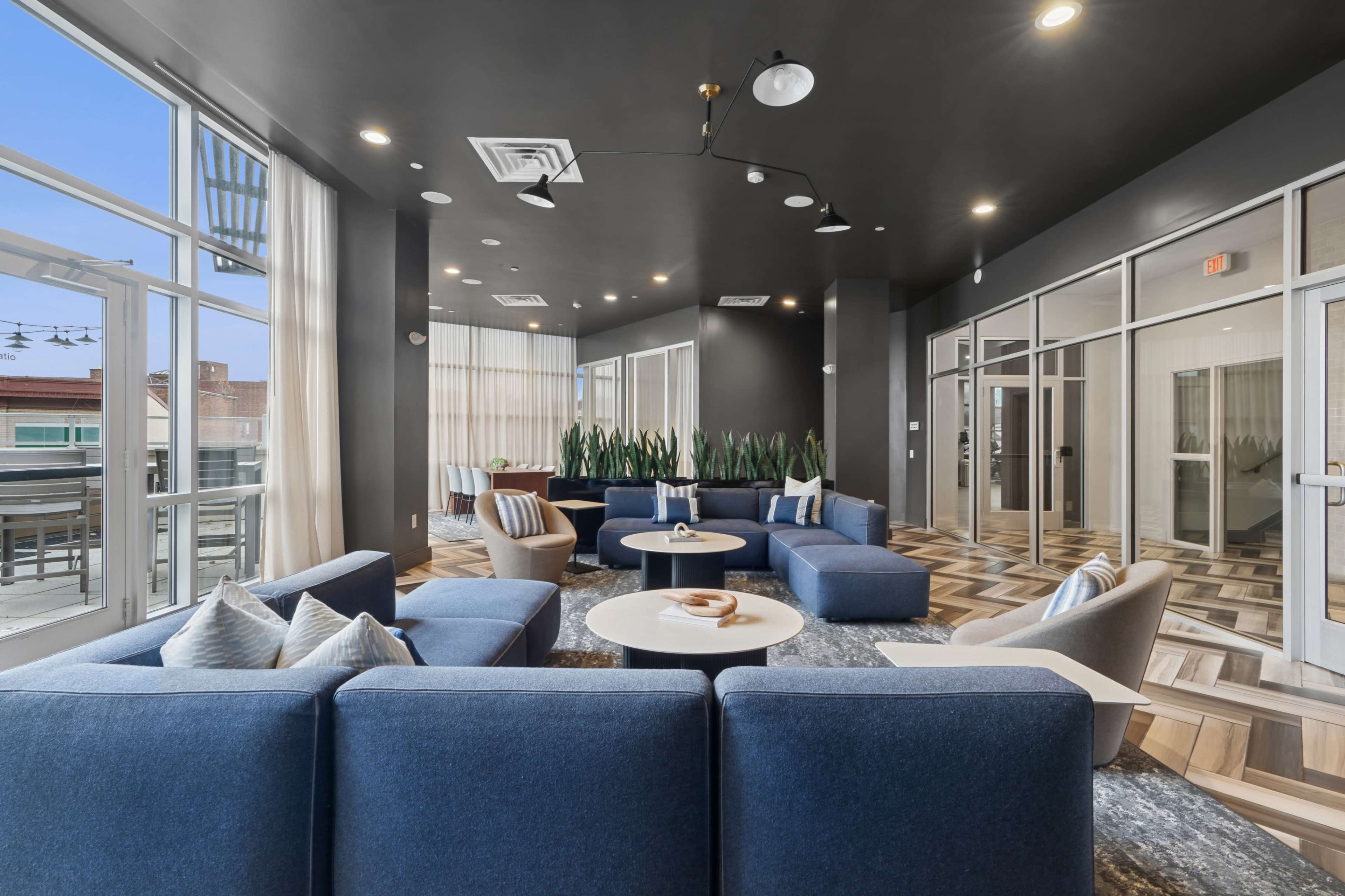 The image shows a modern lounge area featuring blue sofas, a round coffee table, and tall plants, with large windows allowing natural light to enter.