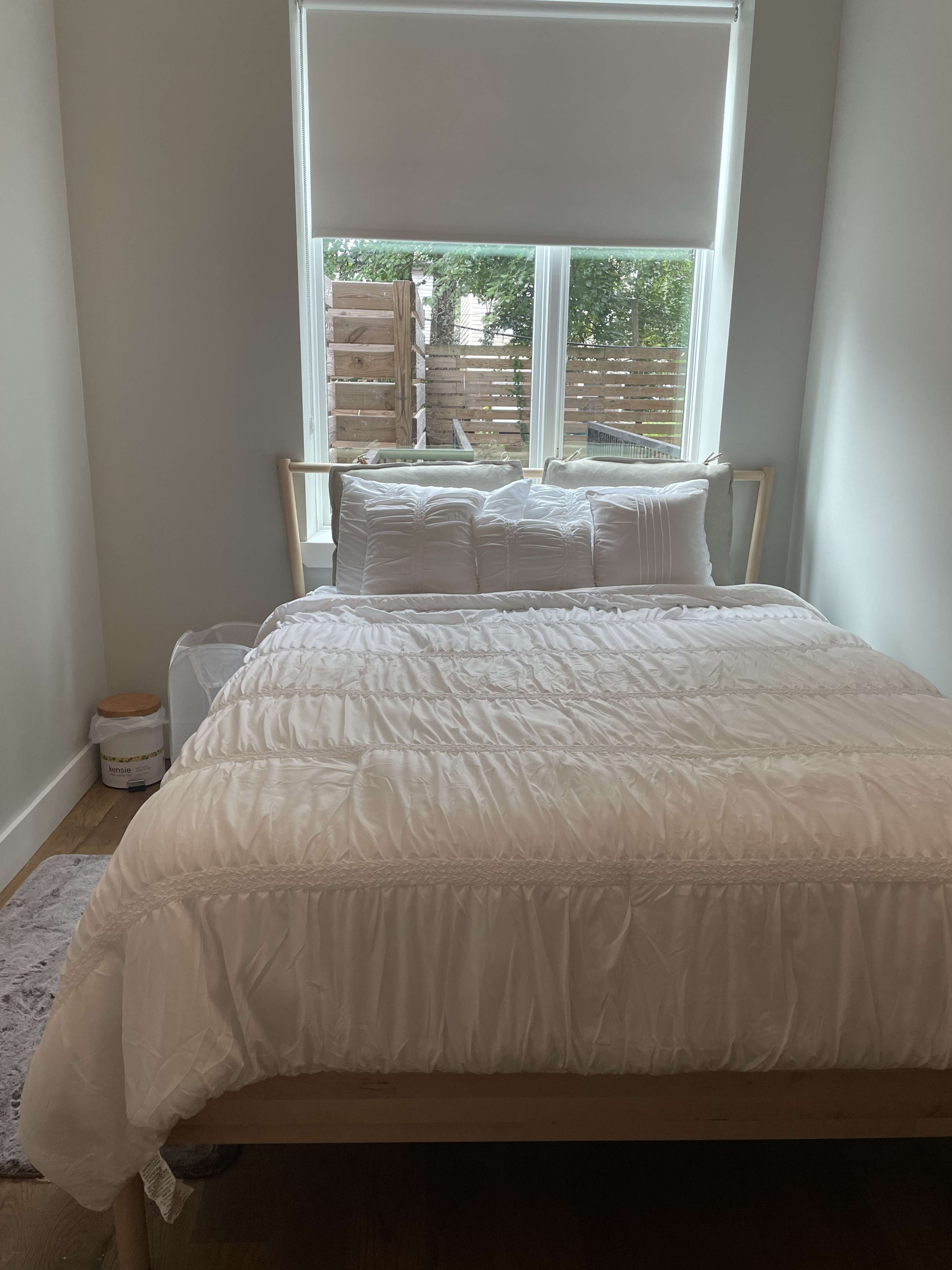 A neatly made bed with a white comforter and pillows in a well-lit bedroom with a window overlooking a wooden patio.
