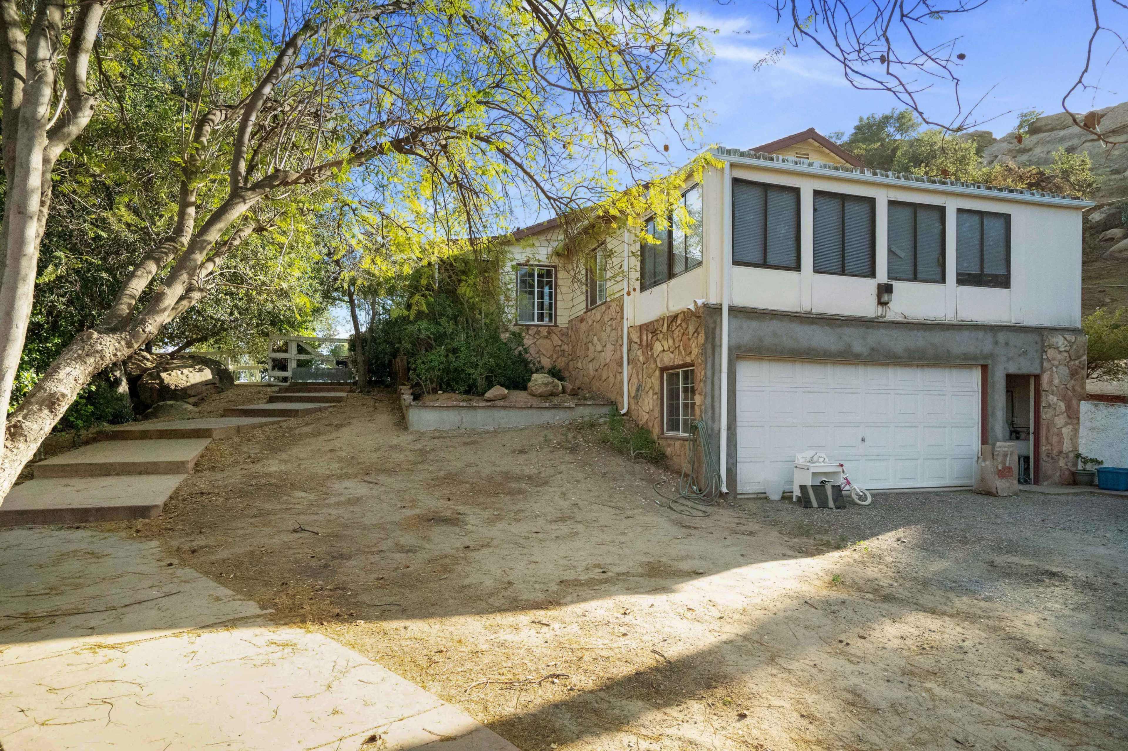 A two-story house with a stone exterior features a garage and is surrounded by trees and a gravel driveway.