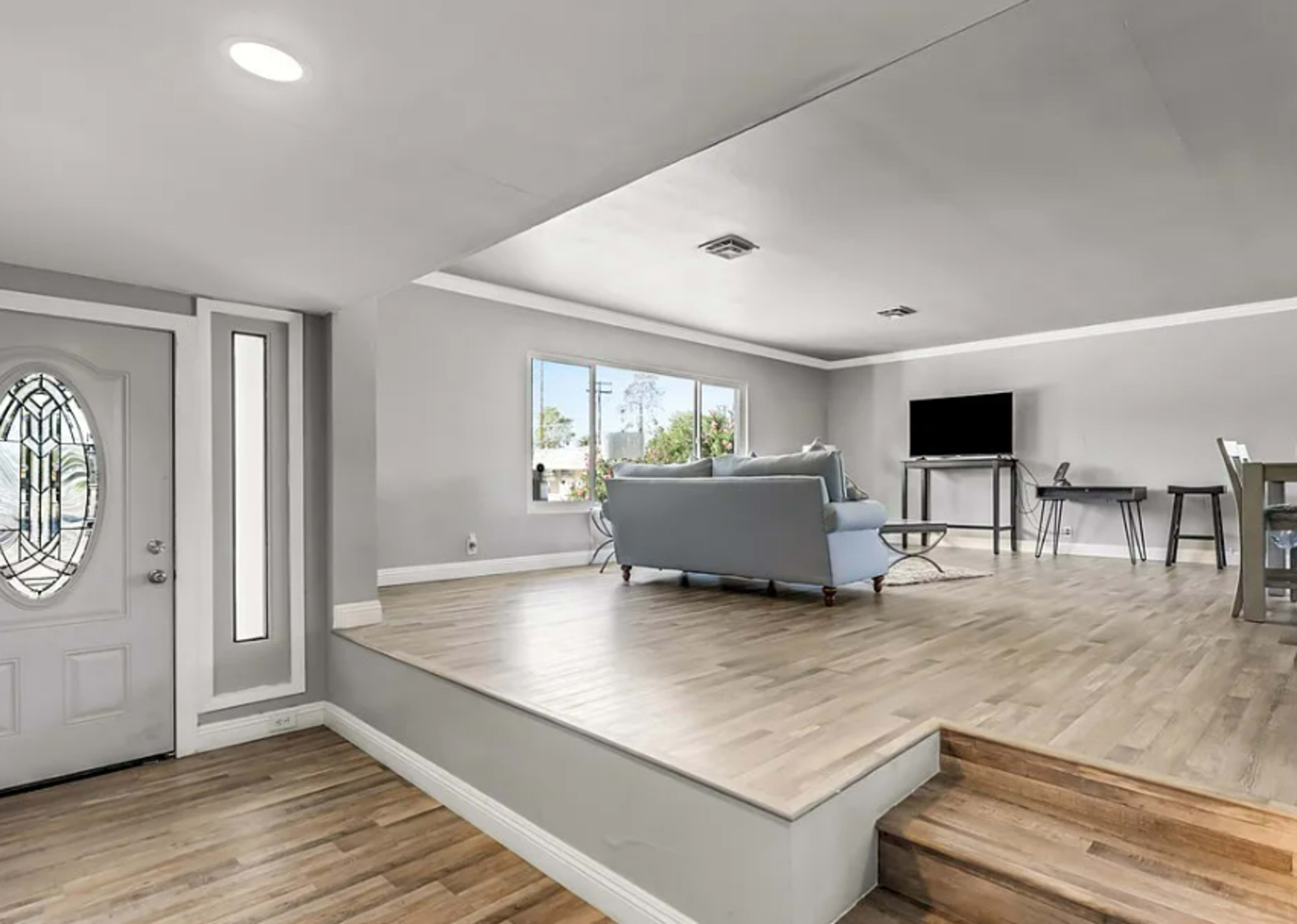 The image shows a modern living space with a gray color scheme, featuring a light-colored sofa, a television, and a set of stairs leading to a raised area near the entrance.
