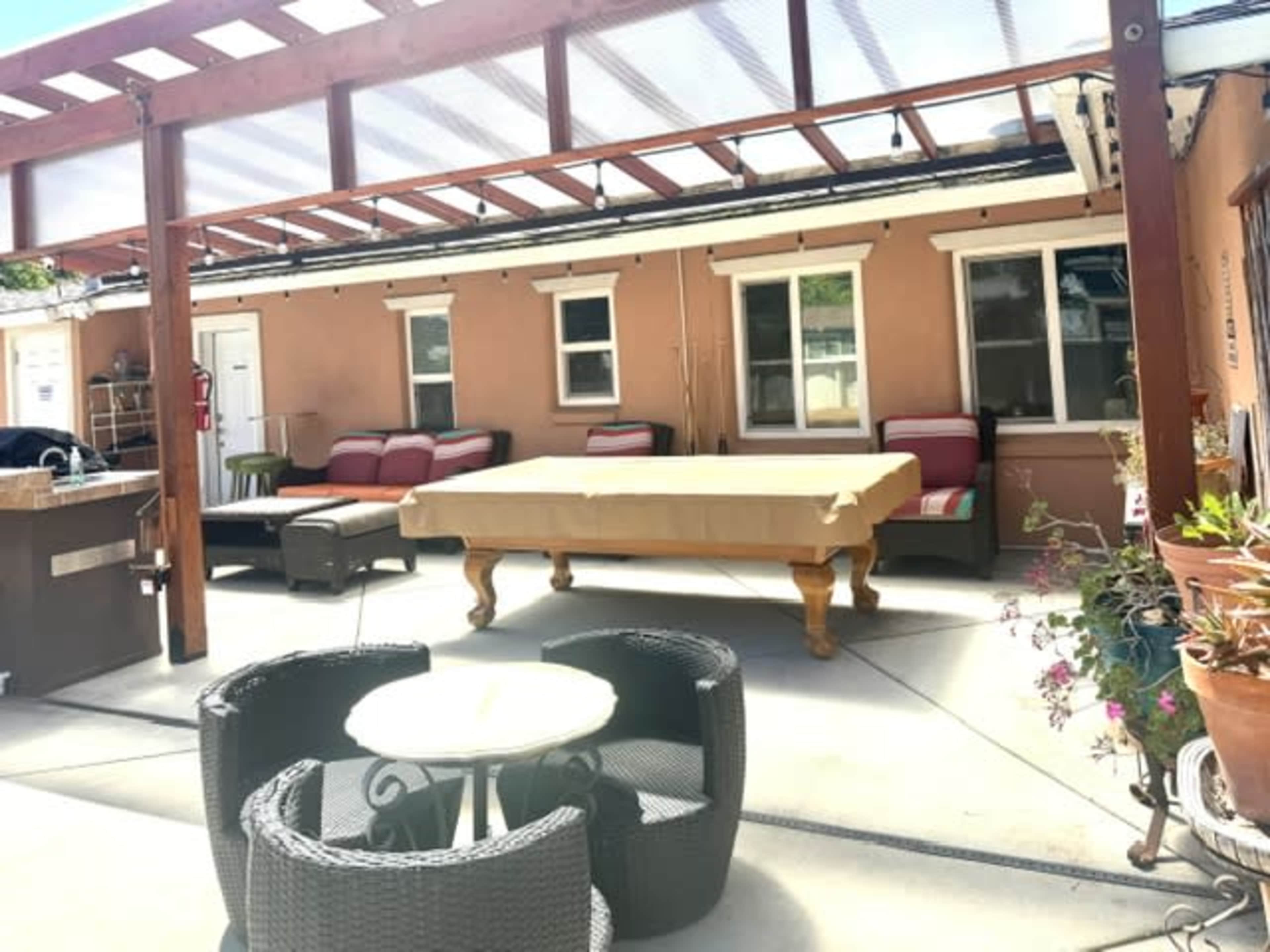 The image shows a patio area with a pool table, outdoor seating, and potted plants under a wooden pergola.