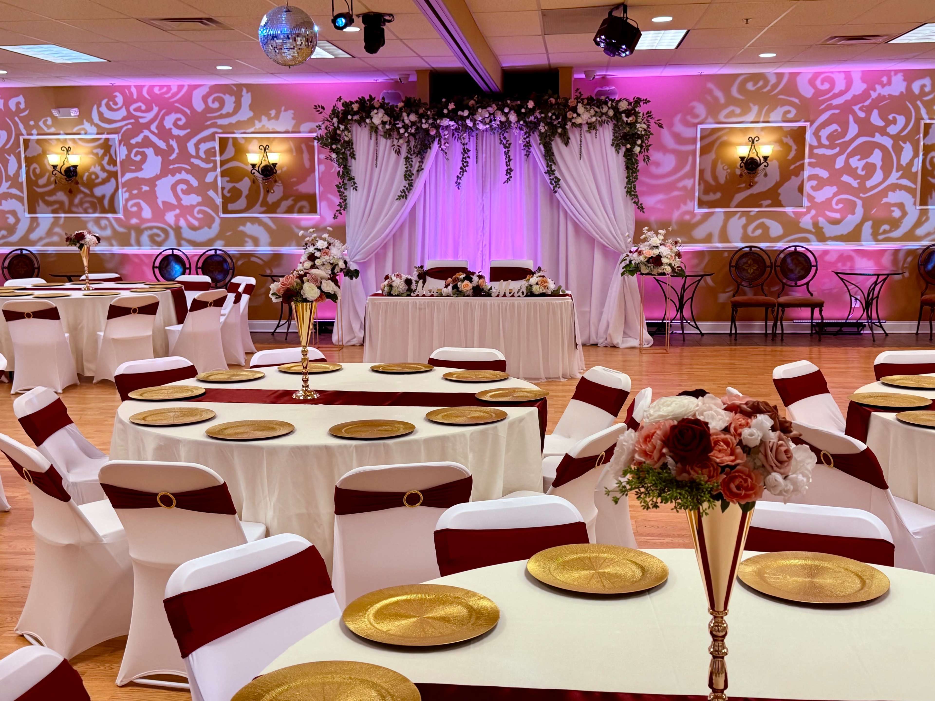 The banquet hall features elegantly set tables with gold chargers and white tablecloths, adorned with red sashes, under decorative lighting and floral arrangements.