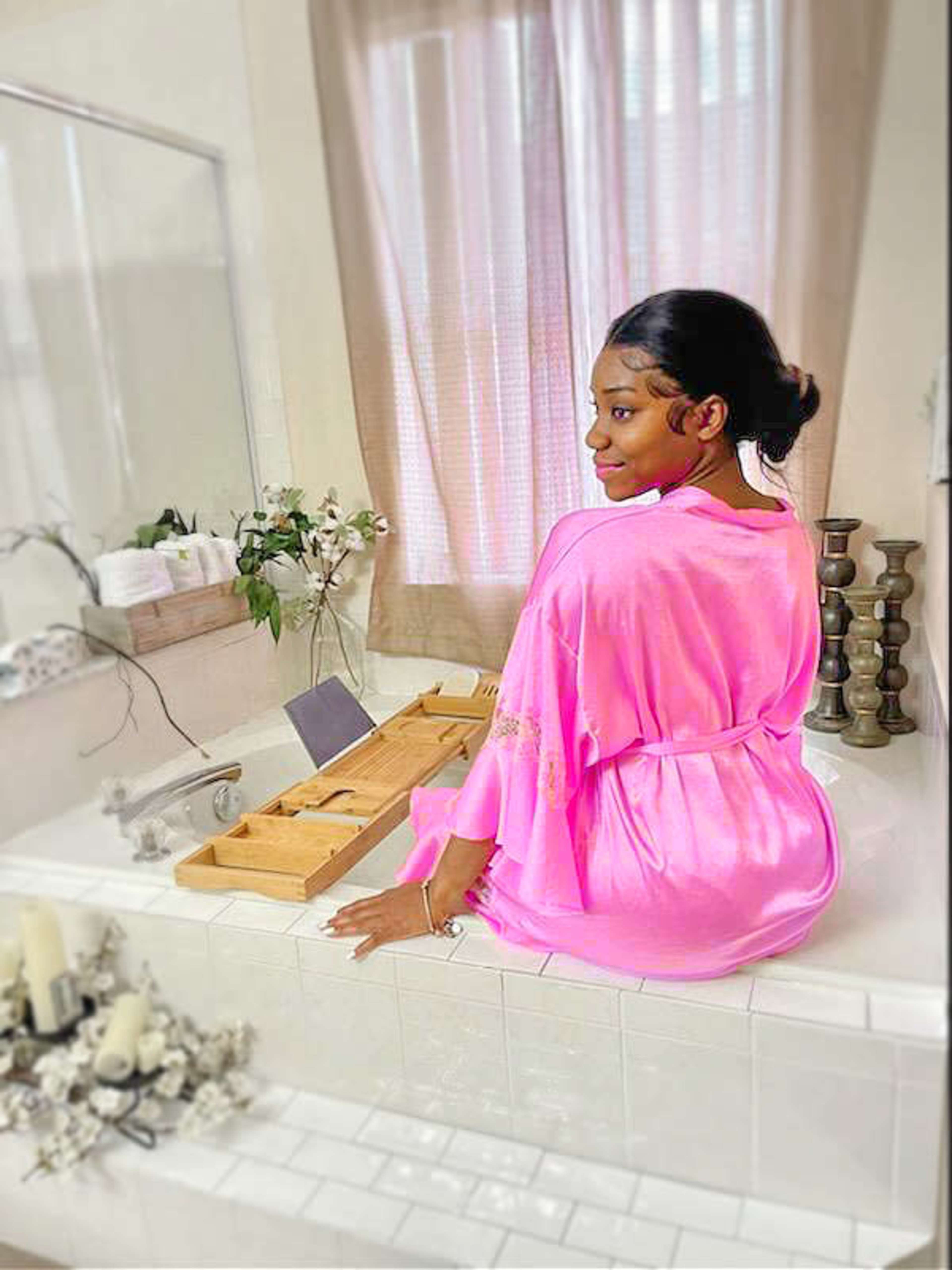 A woman in a pink robe sits on the edge of a bathtub in a well-lit bathroom.