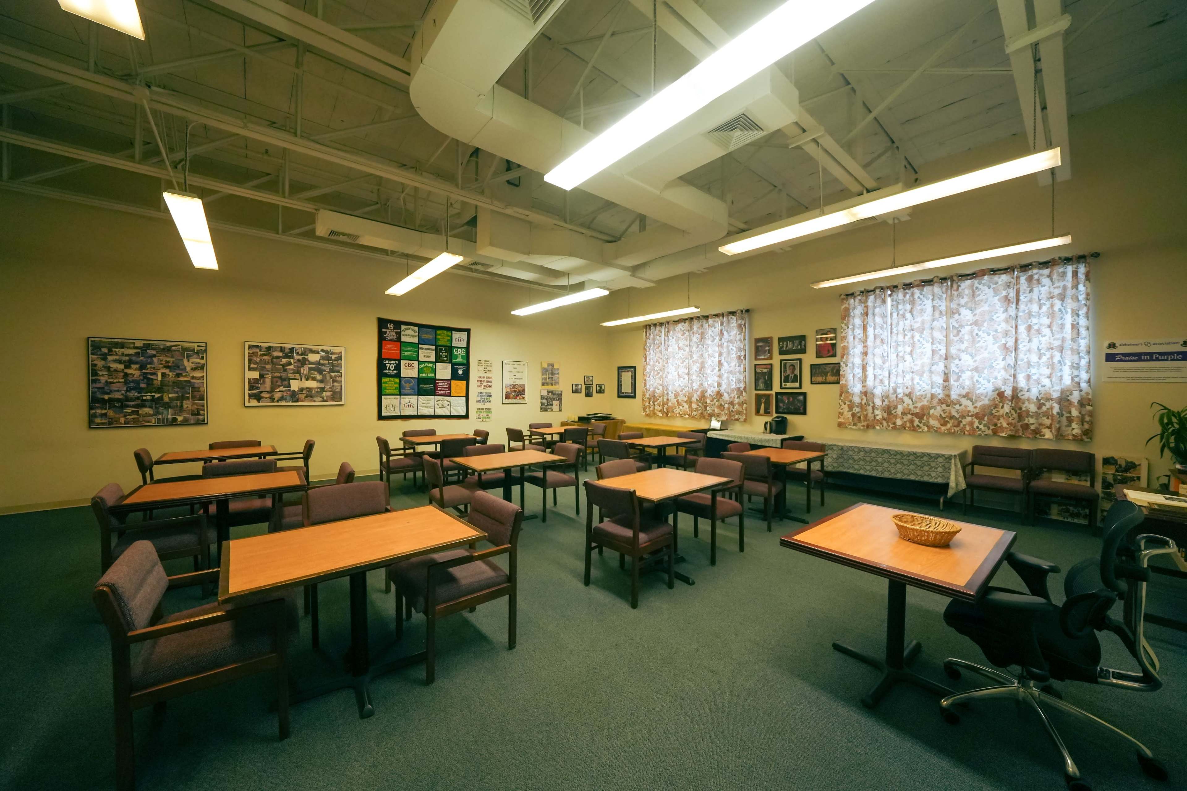 A spacious classroom with several wooden tables and chairs arranged around the room, and large windows covered with floral curtains.